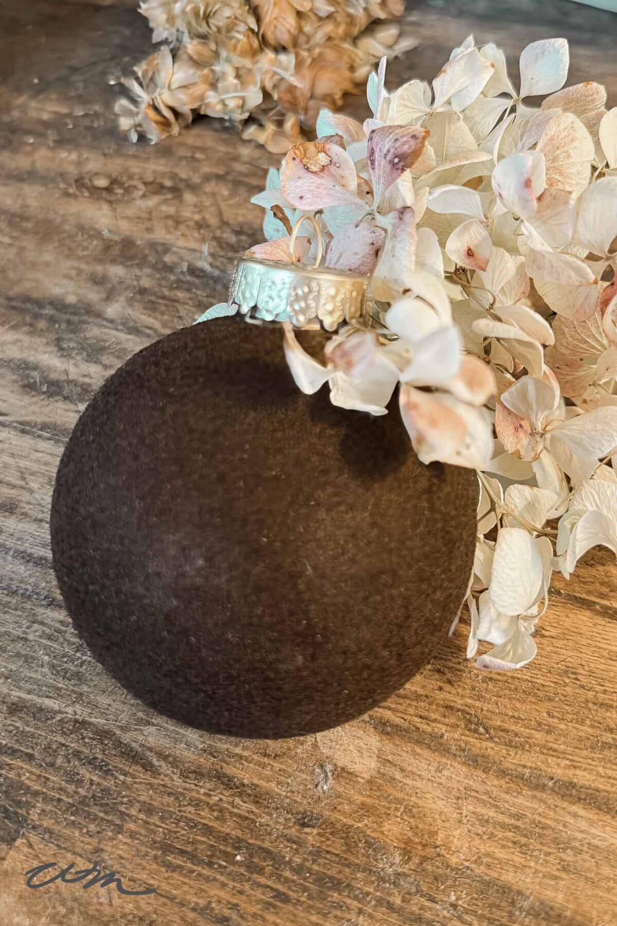 A dark brown, velvet-covered Christmas ornament with a silver cap rests on a wooden surface beside a cluster of dried, pale hydrangea flowers.