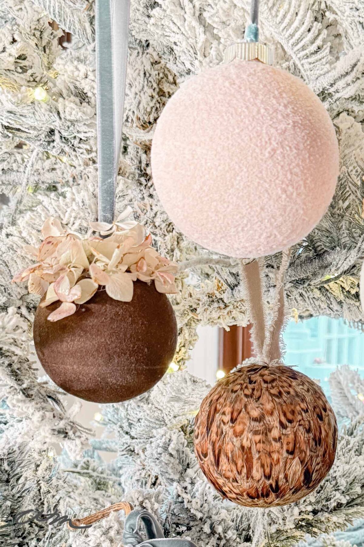 Three Christmas ornaments hang on a snowy white Christmas tree: one brown with dried flowers, one fuzzy pale pink, and one brown with a feather-like texture. Warm white lights twinkle in the background.