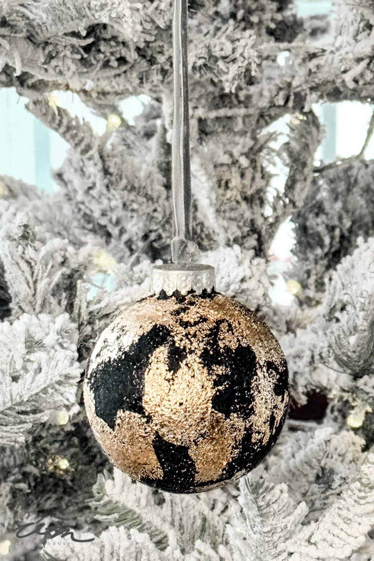 A gold and black Christmas ornament, inspired by DIY Velvet Ornaments, hangs from a snowy, white tree branch. The artificial snow glistens as small white lights twinkle softly in the background.