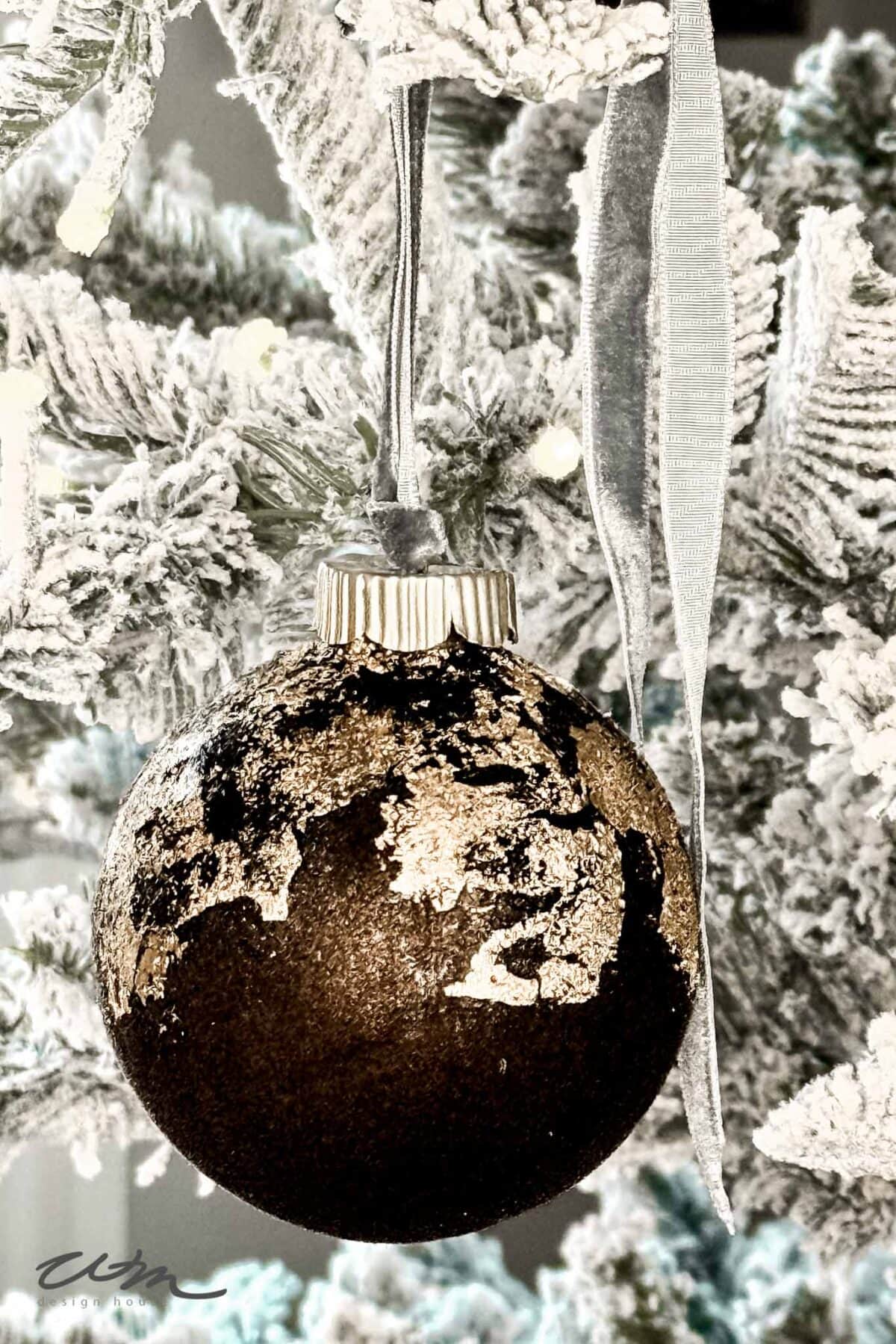 A close-up of a rustic, dark DIY Velvet Ornament with gold leaf patches hangs from a frosted, snow-covered tree branch, decorated with a silver ribbon.