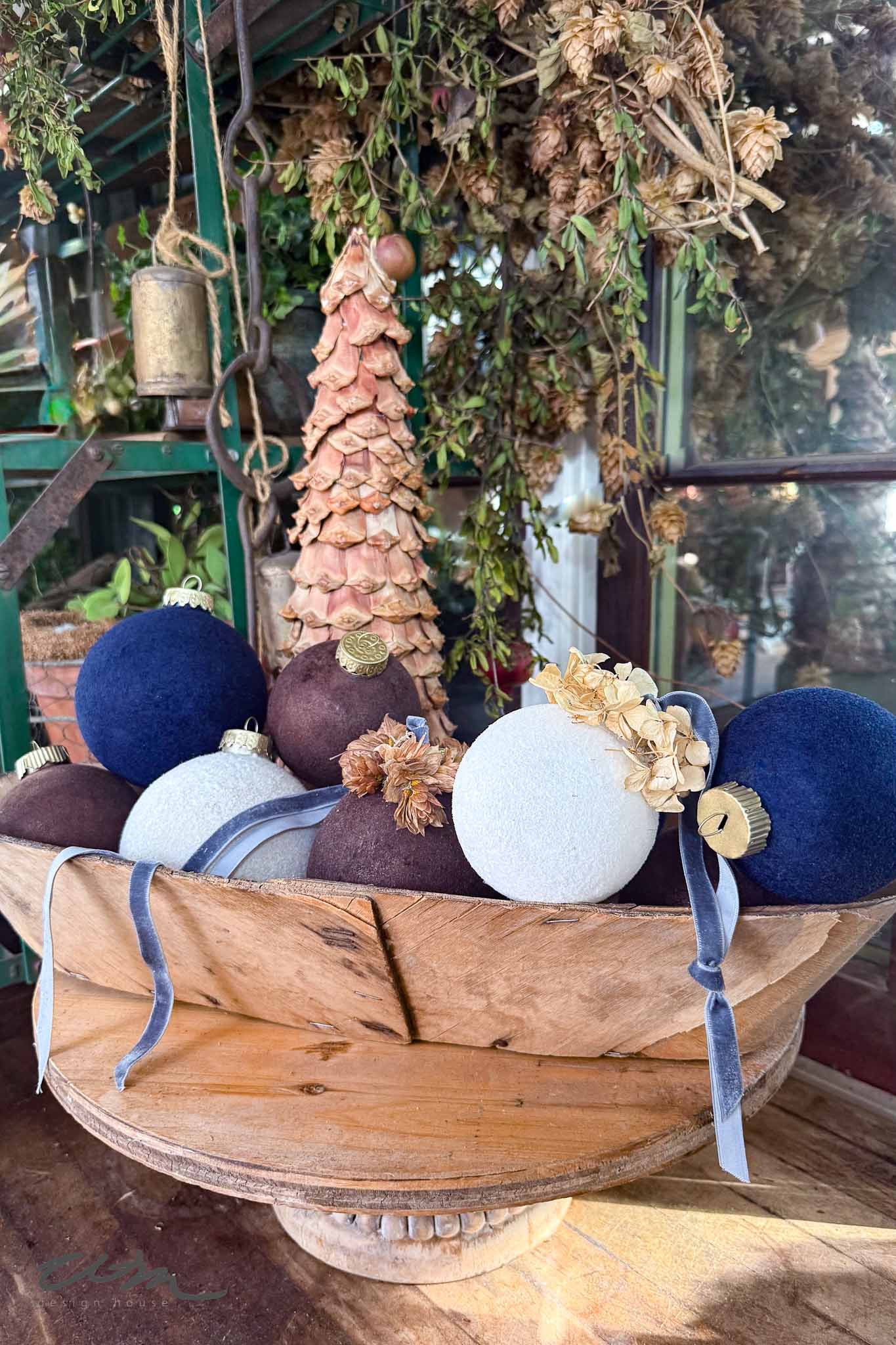 A wooden bowl holds blue, brown, and white DIY Velvet Ornaments with ribbons and dried flower accents. In the background, a pinecone tree and dried hanging plants are visible.