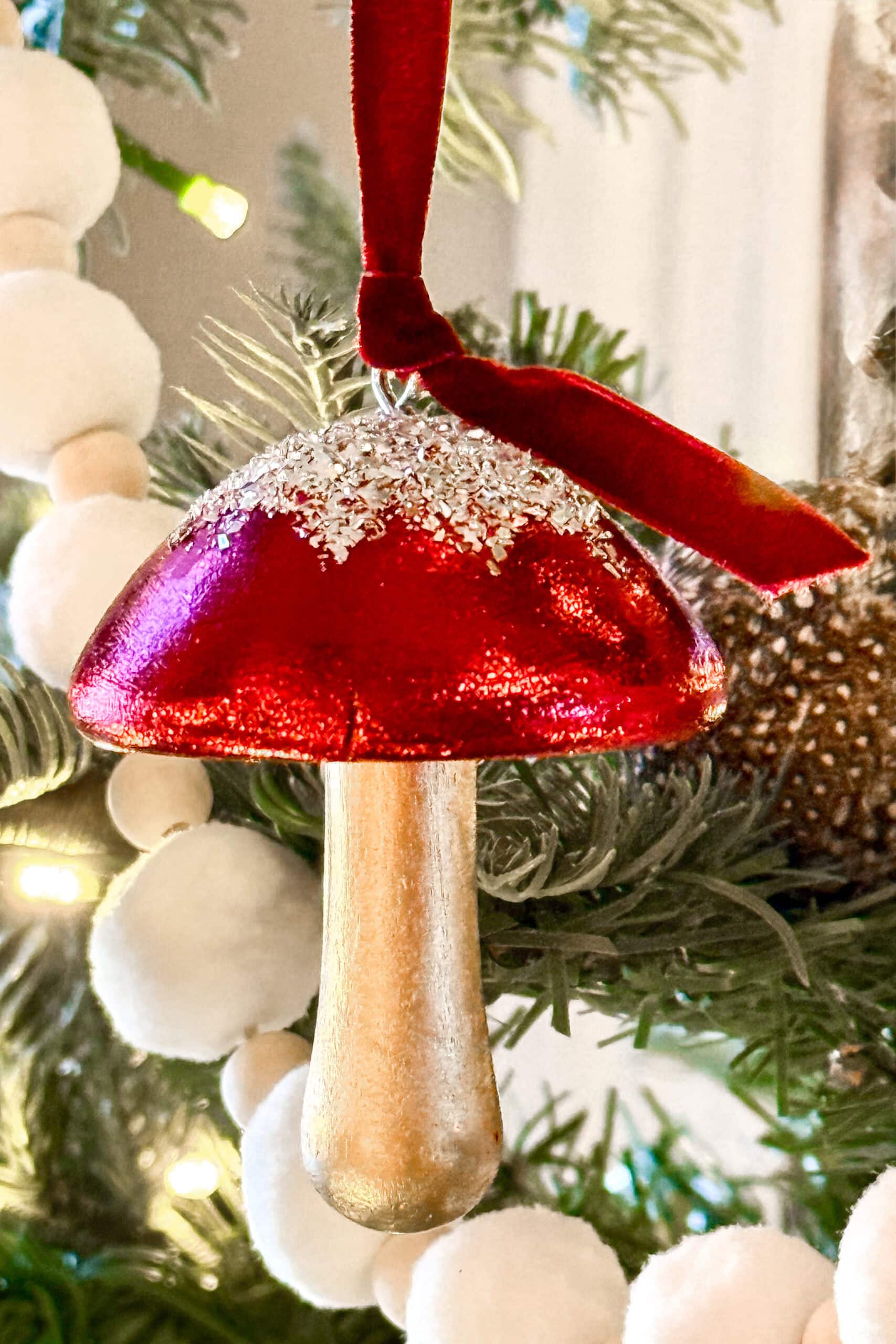 A shiny red and gold mushroom ornament with glitter and a red velvet ribbon hangs from a Christmas tree branch decorated with white pom-poms.