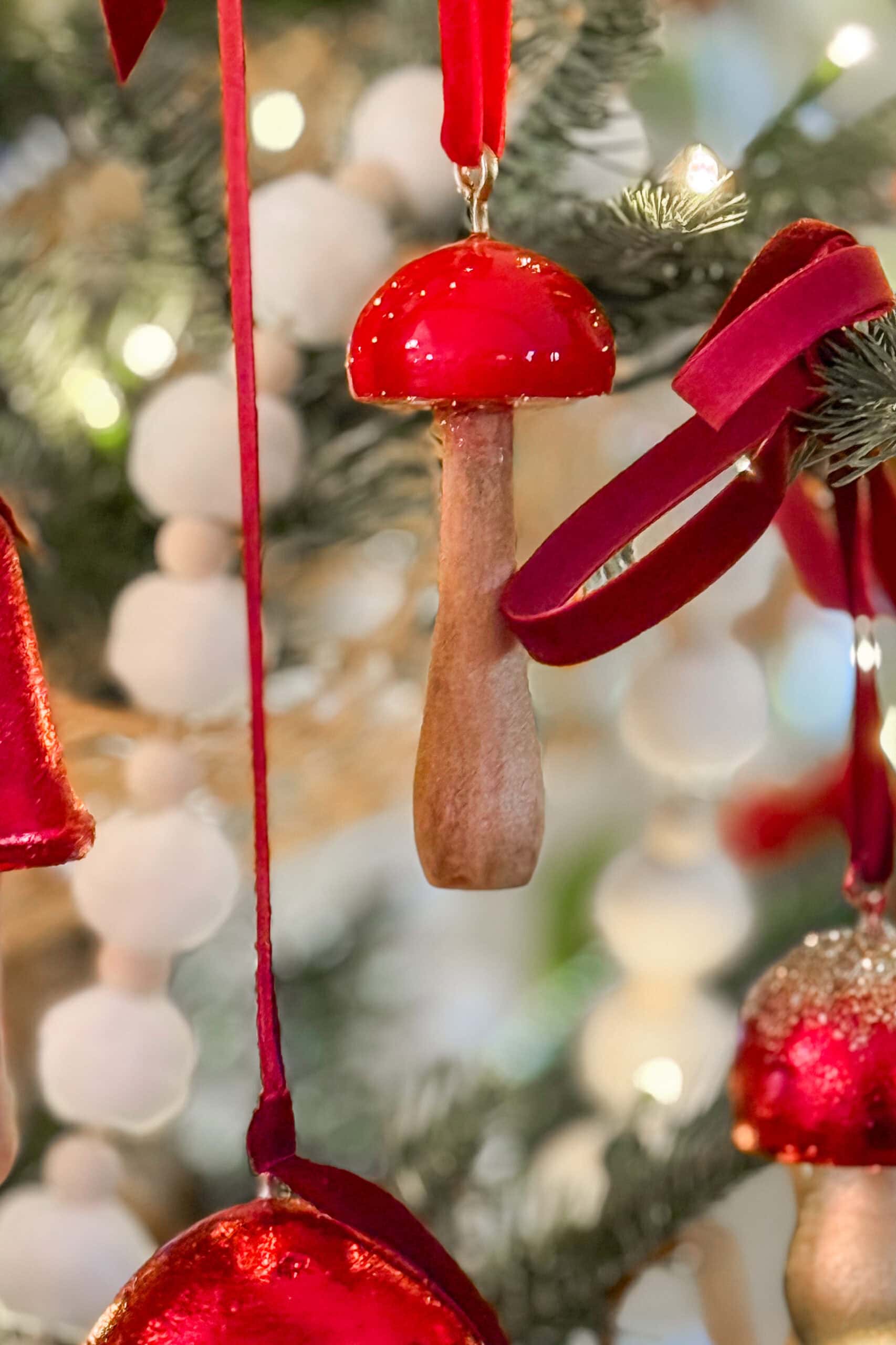 A close-up of a Christmas tree ornament shaped like a red and beige mushroom, hanging among red velvet ribbons and white garland on a festive tree.