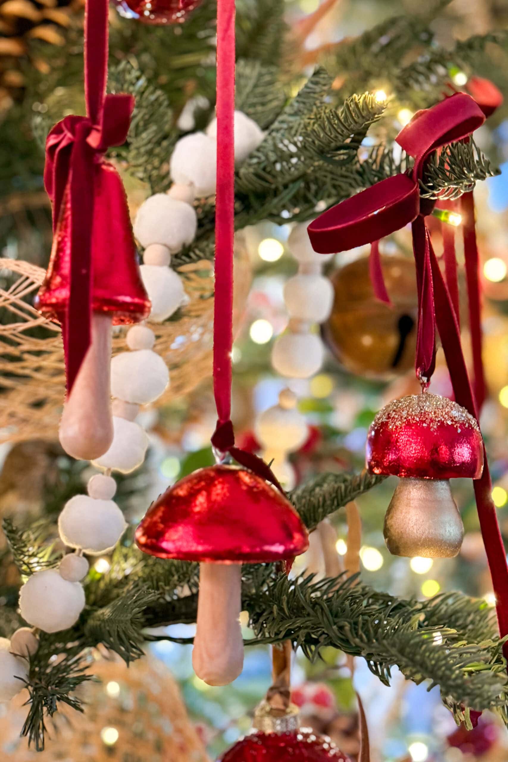 Red and white mushroom-shaped ornaments hang from a Christmas tree, decorated with velvet ribbons and white pompom garlands, surrounded by green pine needles and festive lights.