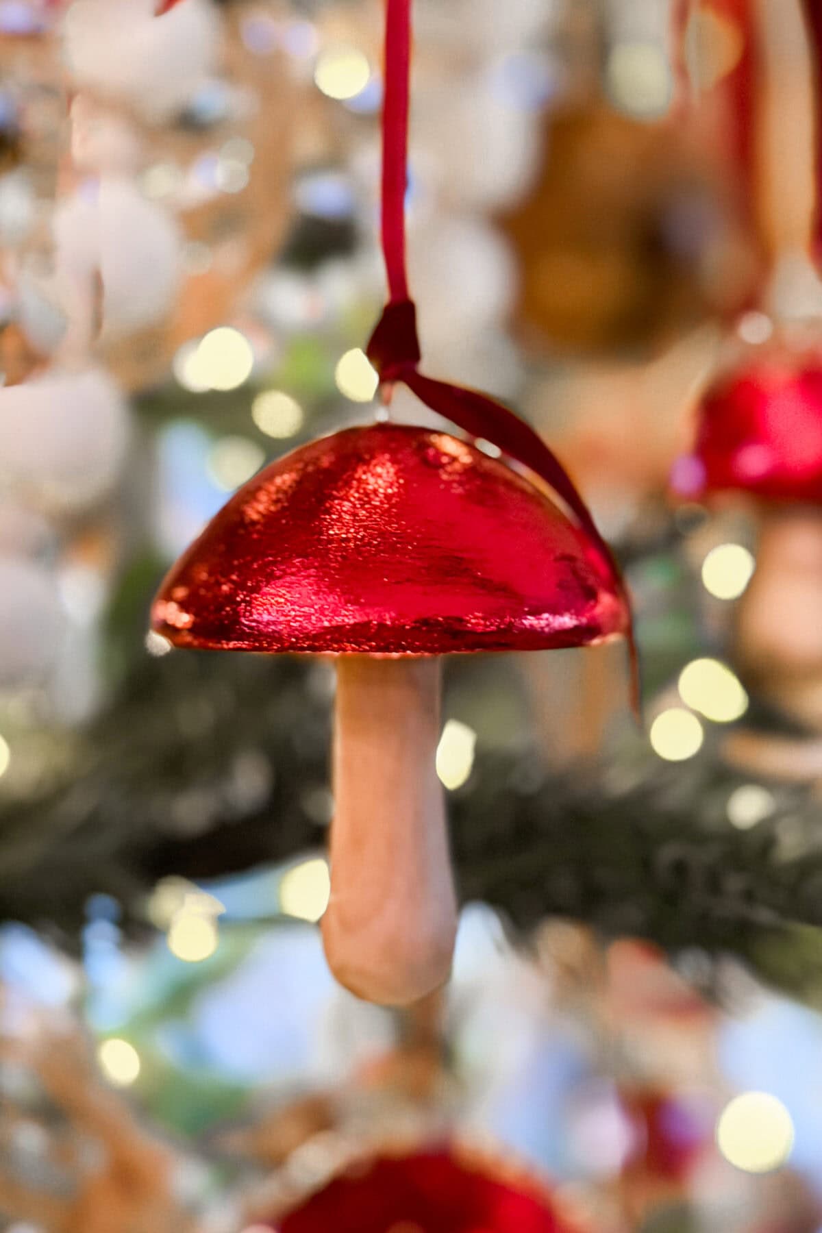 A shiny red mushroom-shaped ornament with a wooden stem hangs from a red ribbon on a Christmas tree, with blurred lights and decorations in the background.
