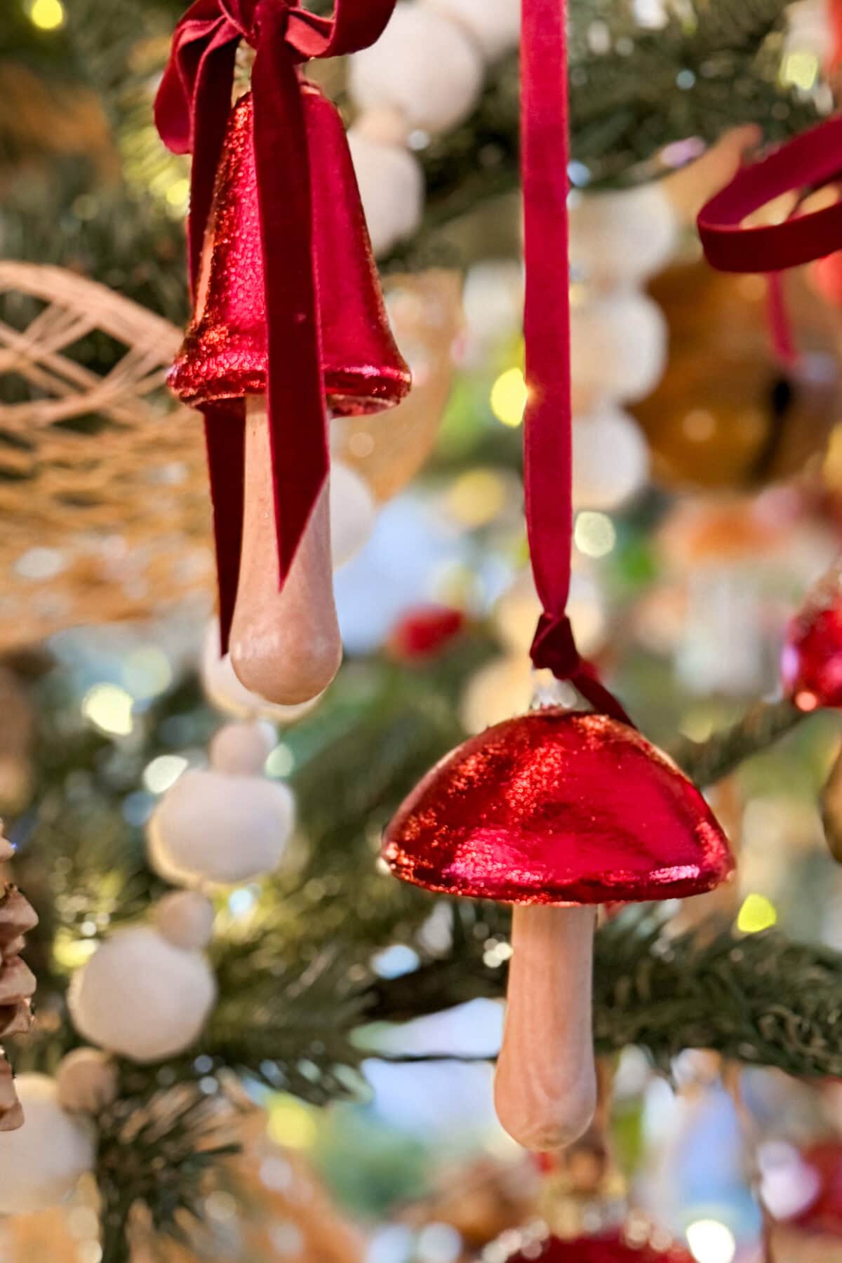 Two Christmas tree ornaments shaped like mushrooms with shiny red caps and beige stems hang on a pine branch, decorated with red ribbons and surrounded by blurred festive lights and decorations in the background.