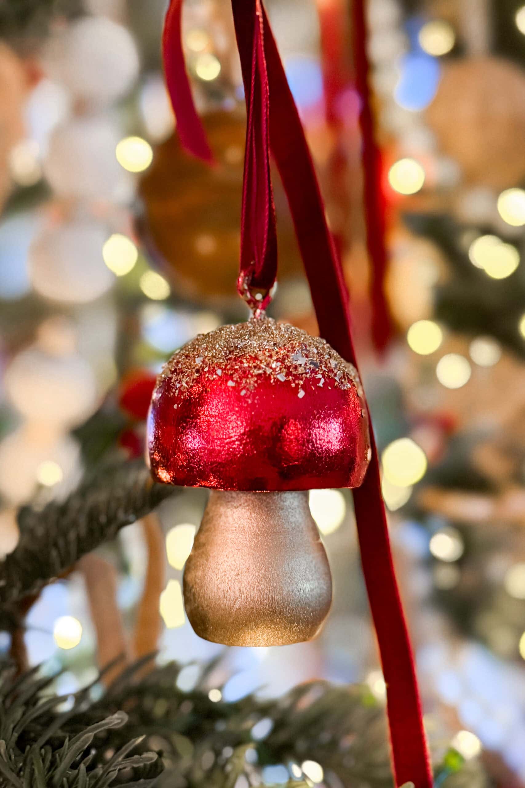 A glittery red and gold mushroom-shaped ornament hangs from a red ribbon on a Christmas tree, with festive blurred lights in the background.