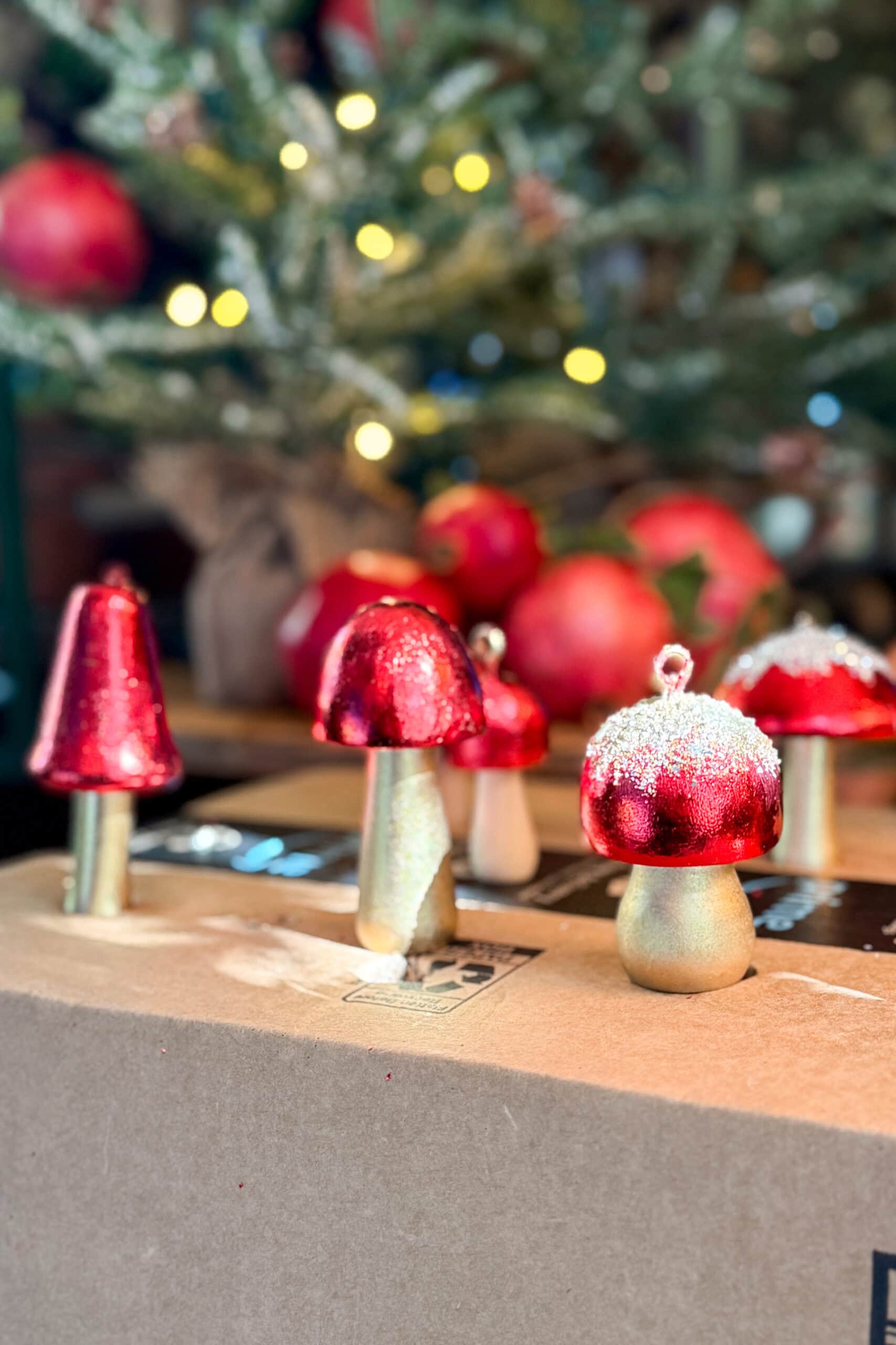 Red and gold glittery mushroom ornaments are displayed on a cardboard box with a decorated Christmas tree and glowing lights in the blurred background.