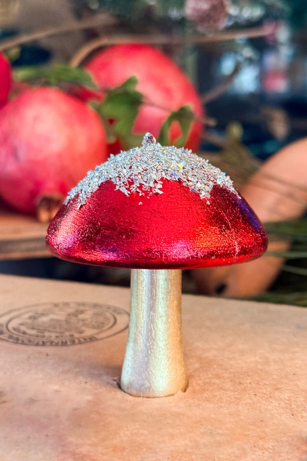 A decorative mushroom with a shiny red cap and sparkling glitter on top, standing on a gold-colored stem. Blurred pomegranates and greenery are visible in the background.
