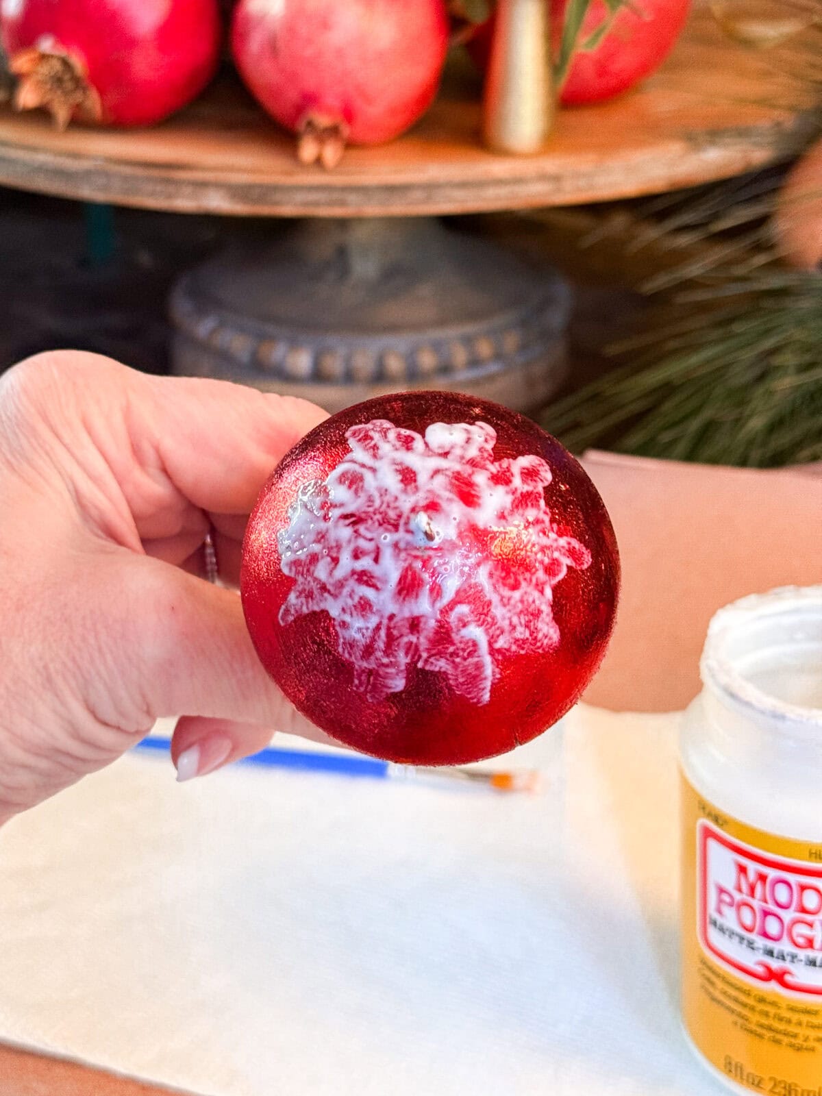 A hand holds a red Christmas ornament partially covered in white glue or Mod Podge, with a jar of Mod Podge and a paintbrush on a napkin below. Pomegranates are visible in the blurred background.