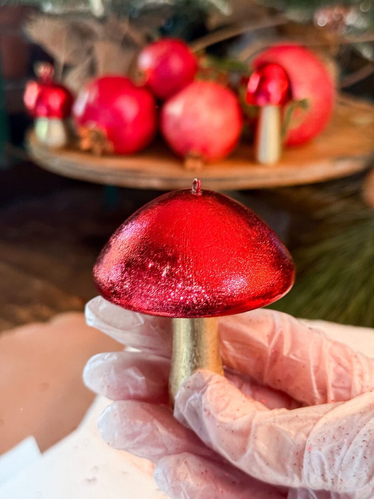 A gloved hand holds a shiny red and gold mushroom-shaped ornament, with red pomegranates and more similar ornaments blurred in the background.