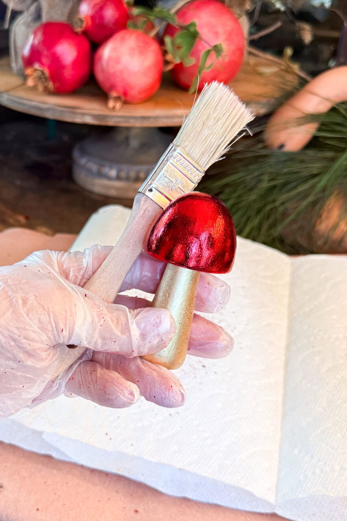 A gloved hand holds a small decorative mushroom, painted red and gold, next to a paintbrush. Pomegranates and greenery are in the blurred background, with a white paper towel underneath.