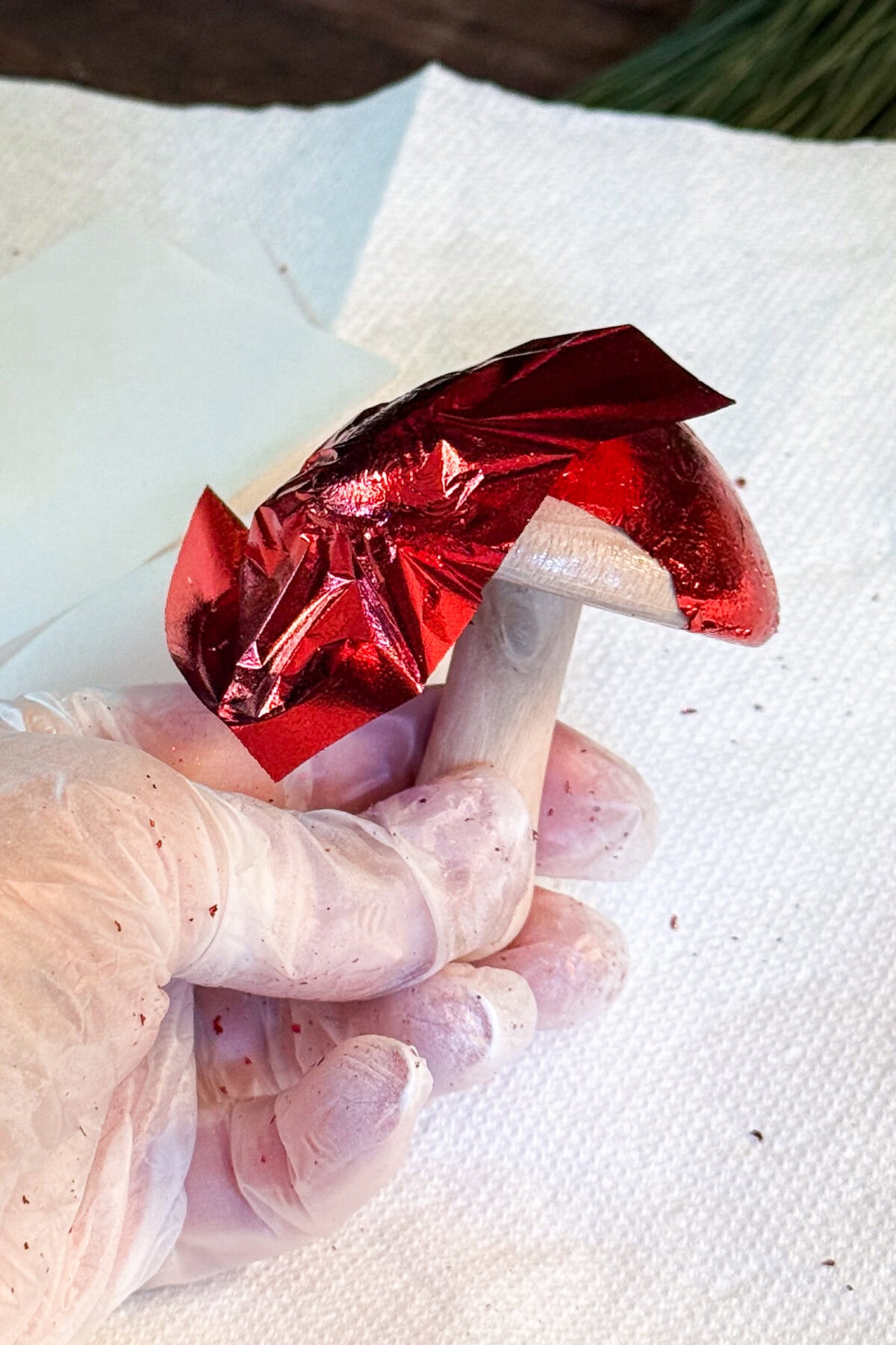 A gloved hand holds a mushroom while wrapping its cap in shiny red foil, with a white paper towel underneath.