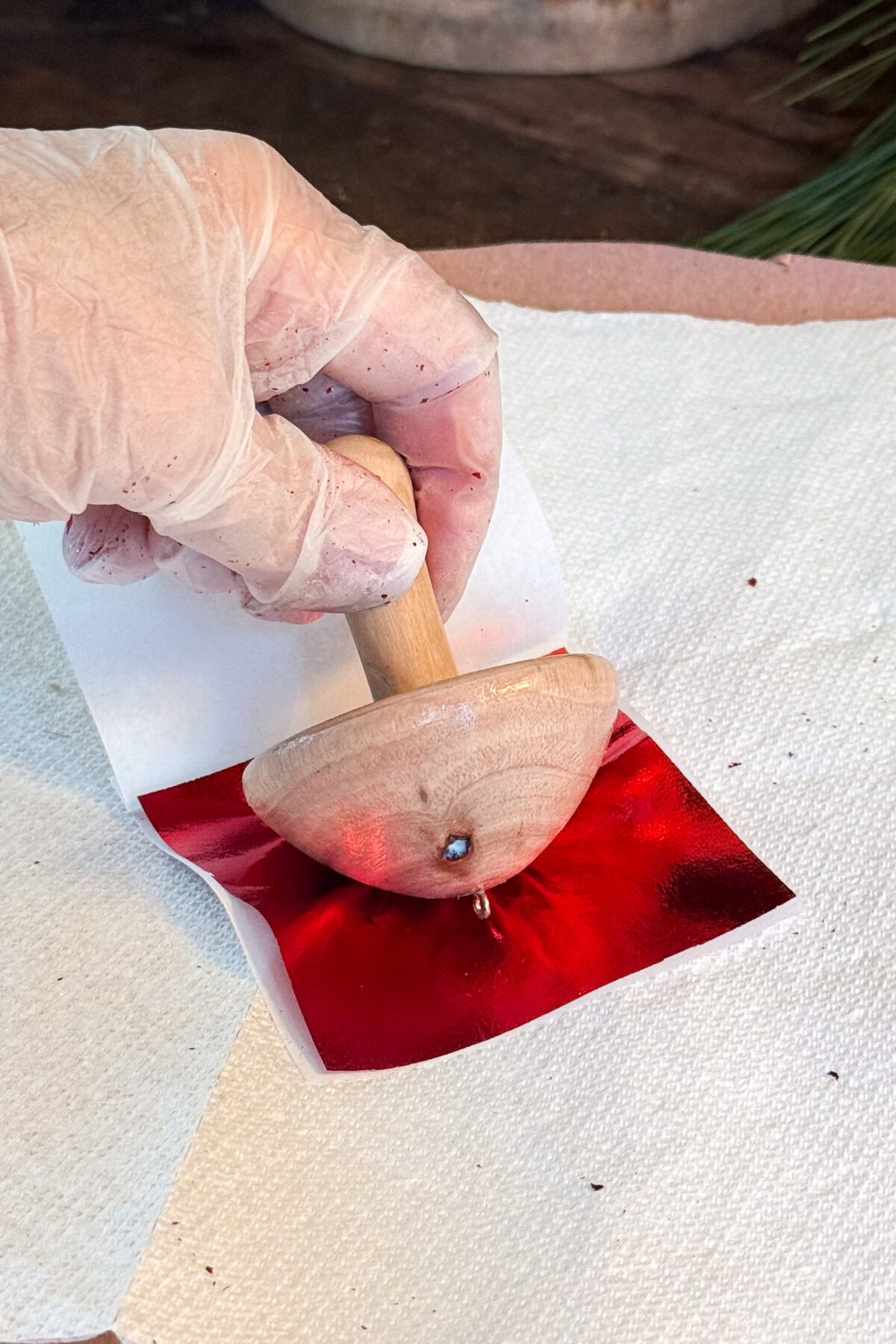 A gloved hand holds a wooden spinning top pressing down on a small square of shiny red foil placed on a white paper towel.