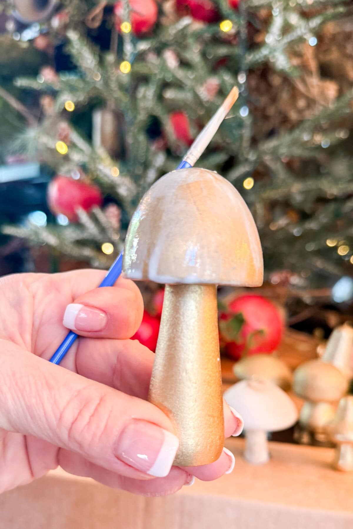 A hand with a French manicure holds a small paintbrush and a gold-painted mushroom ornament, with a decorated Christmas tree and more ornaments blurred in the background.