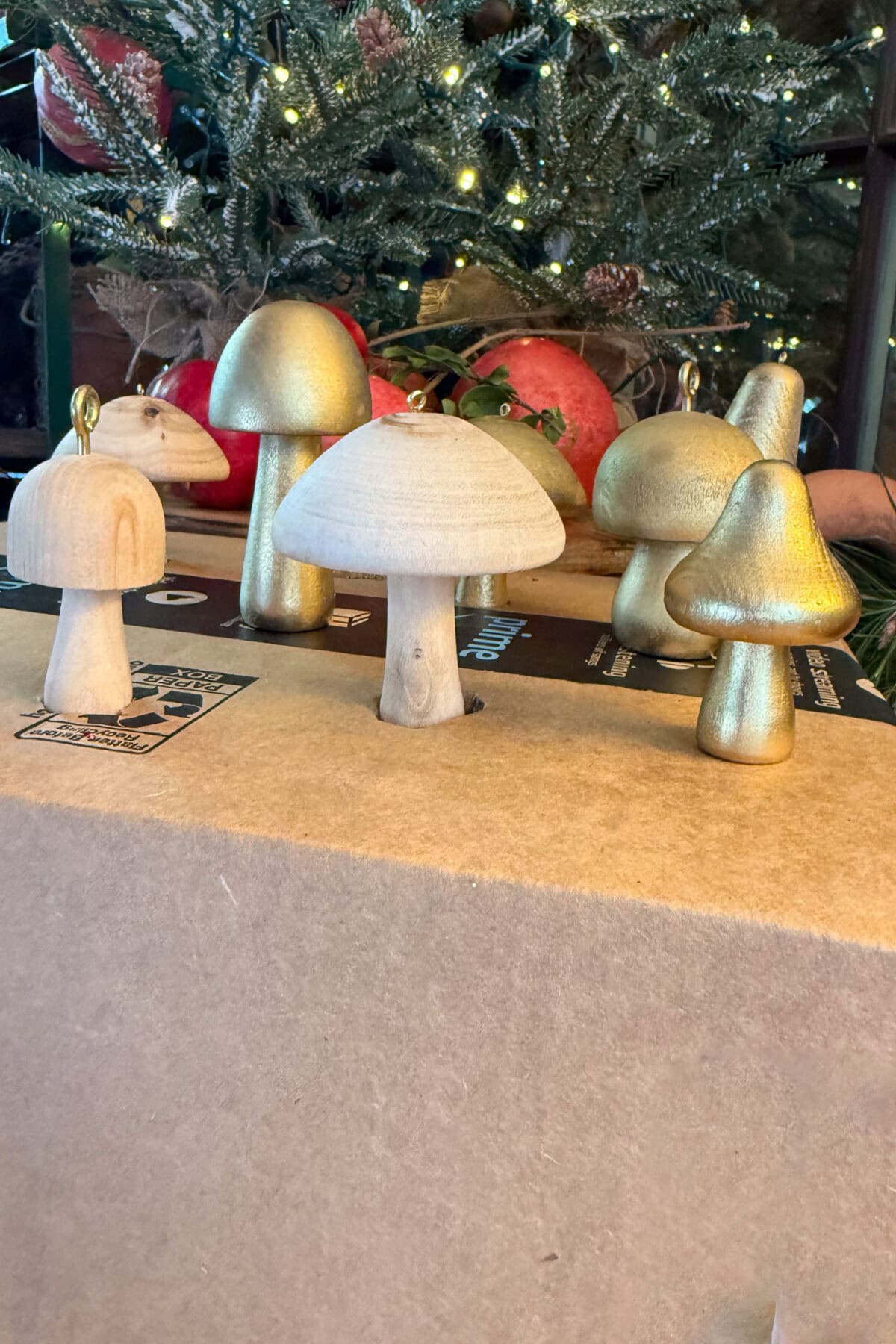 A group of wooden and gold-painted mushroom ornaments are displayed on a beige surface in front of a Christmas tree decorated with white lights.