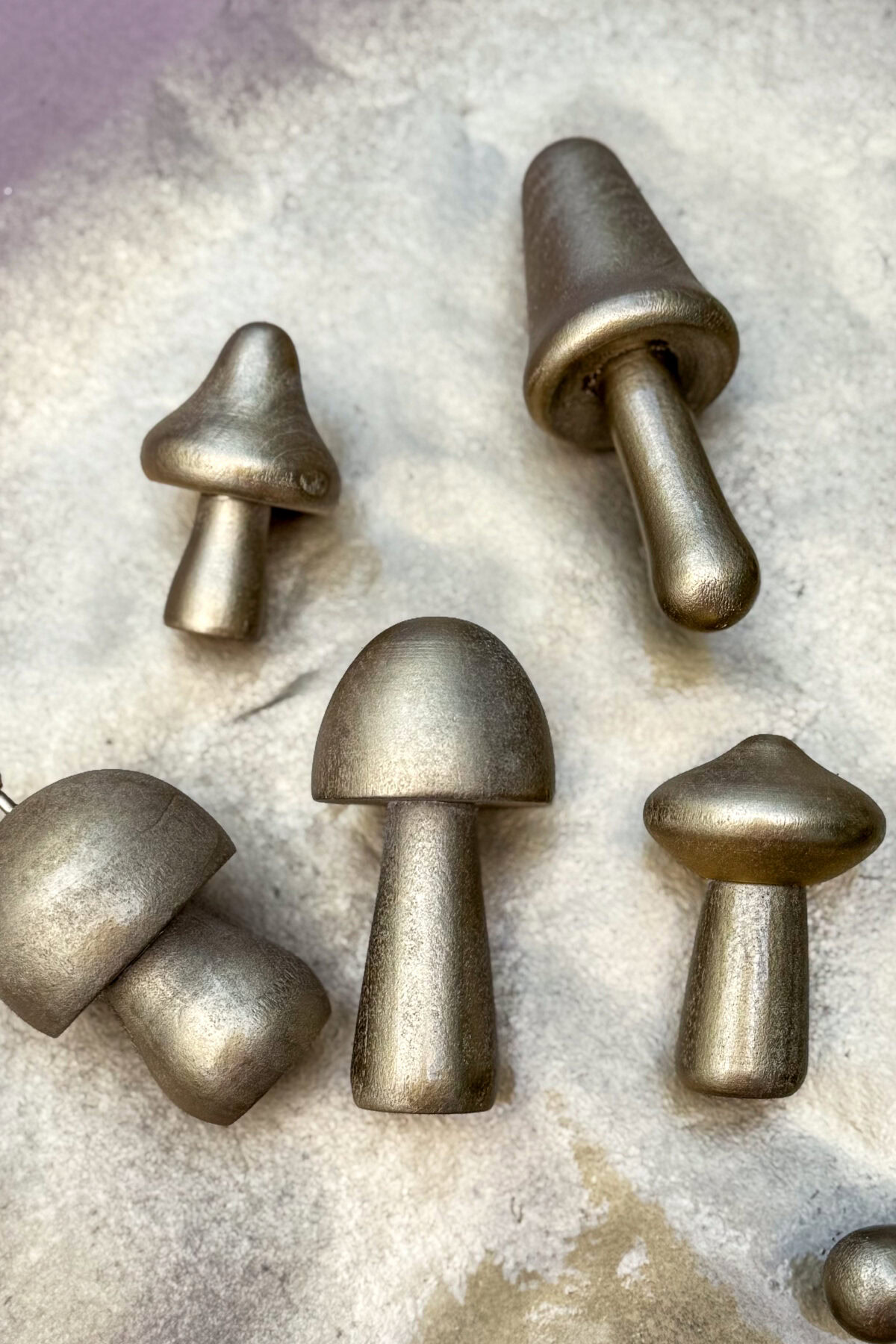 Six metallic, mushroom-shaped objects of varying sizes and shapes are arranged on a textured, light-colored surface. The objects reflect light and have a brushed or matte finish.