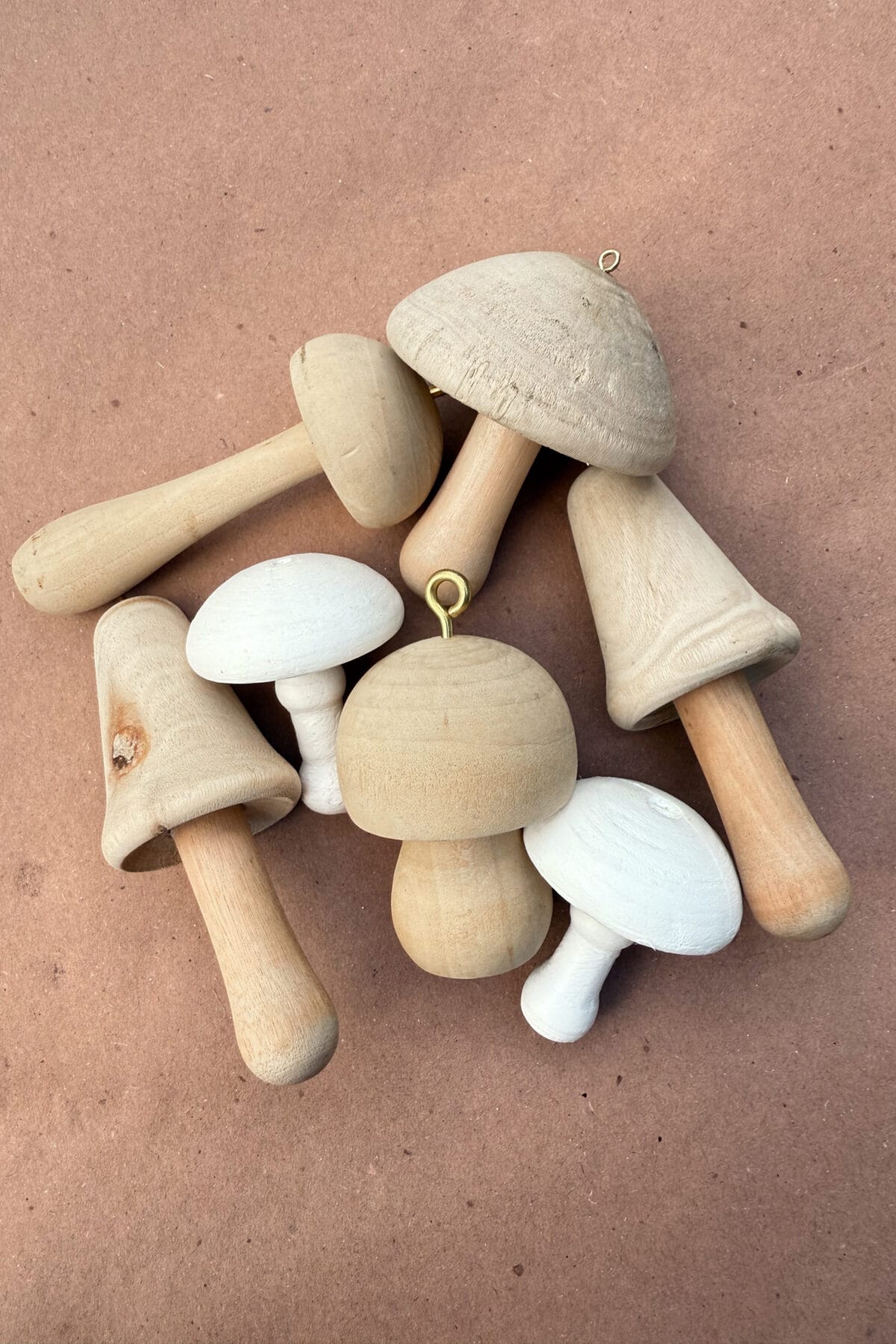 A cluster of various wooden mushrooms with different shapes and sizes is arranged on a brown surface. Some mushrooms are unfinished wood, while others are painted white. Two have small metal hooks on top.