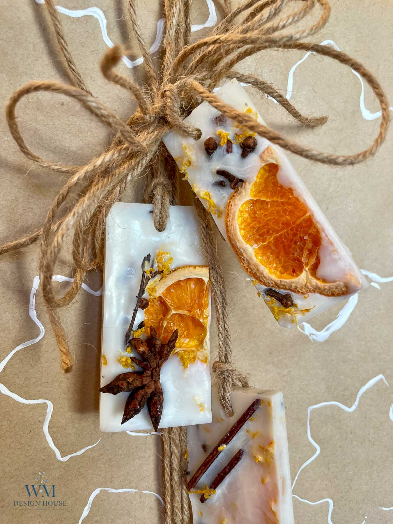 Three rectangular wax melts decorated with dried orange ornaments, whole cloves, and star anise, tied together with twine against a brown paper background.