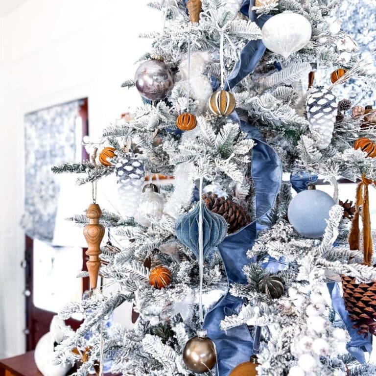 A white flocked Christmas tree, a perfect example of how to decorate a flocked Christmas tree, features blue ribbon, pinecones, orange slices, and blue, silver, and white ornaments in a bright, sunlit room.