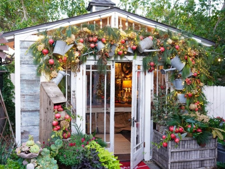A small wooden shed is decorated for the holidays with pine garlands, red ornaments, pomegranate Christmas decor, and metal watering cans. The shed has glass doors, plants in front, and warm lights glowing inside.