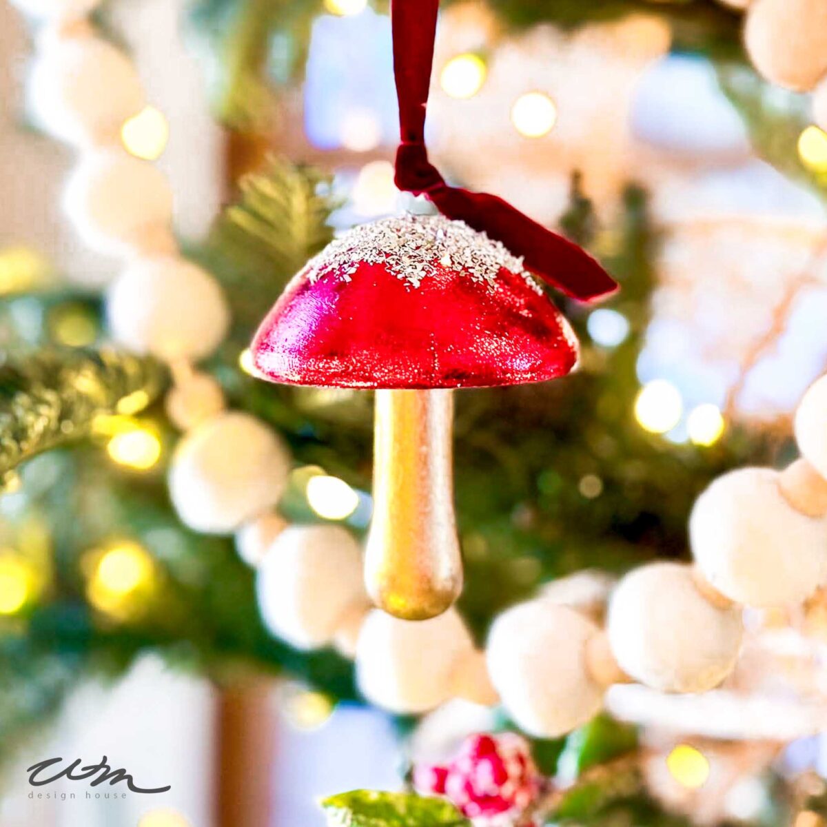 A red and gold wood mushroom ornament with a dusting of glitter hangs from a Christmas tree branch, surrounded by white, round garland decorations.