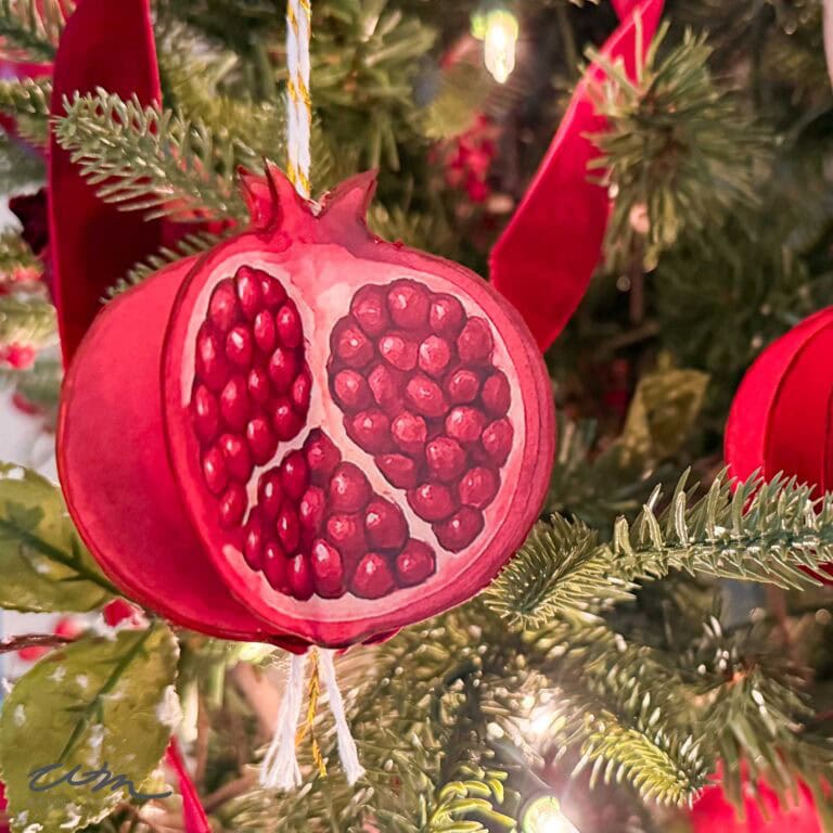 A painted Pomegranate Christmas Ornament, shaped like a pomegranate and revealing detailed red seeds inside, hangs on a Christmas tree adorned with green needles and twinkling lights in the background.