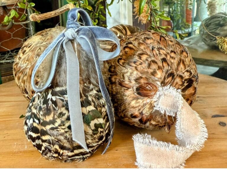 Two DIY Feather Ornaments, covered in patterned feathers, sit on a wooden surface. One is tied with a gray velvet ribbon, the other with a frayed beige fabric ribbon. Greenery appears softly blurred in the background.