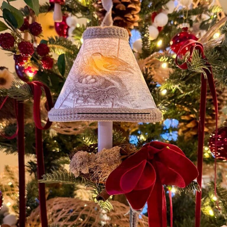 A small lamp with a bird design on the shade, decorated with a red velvet bow, hangs on a Christmas tree adorned with DIY Christmas Tree Candle Lights, twinkling among pinecones and red and white ornaments.