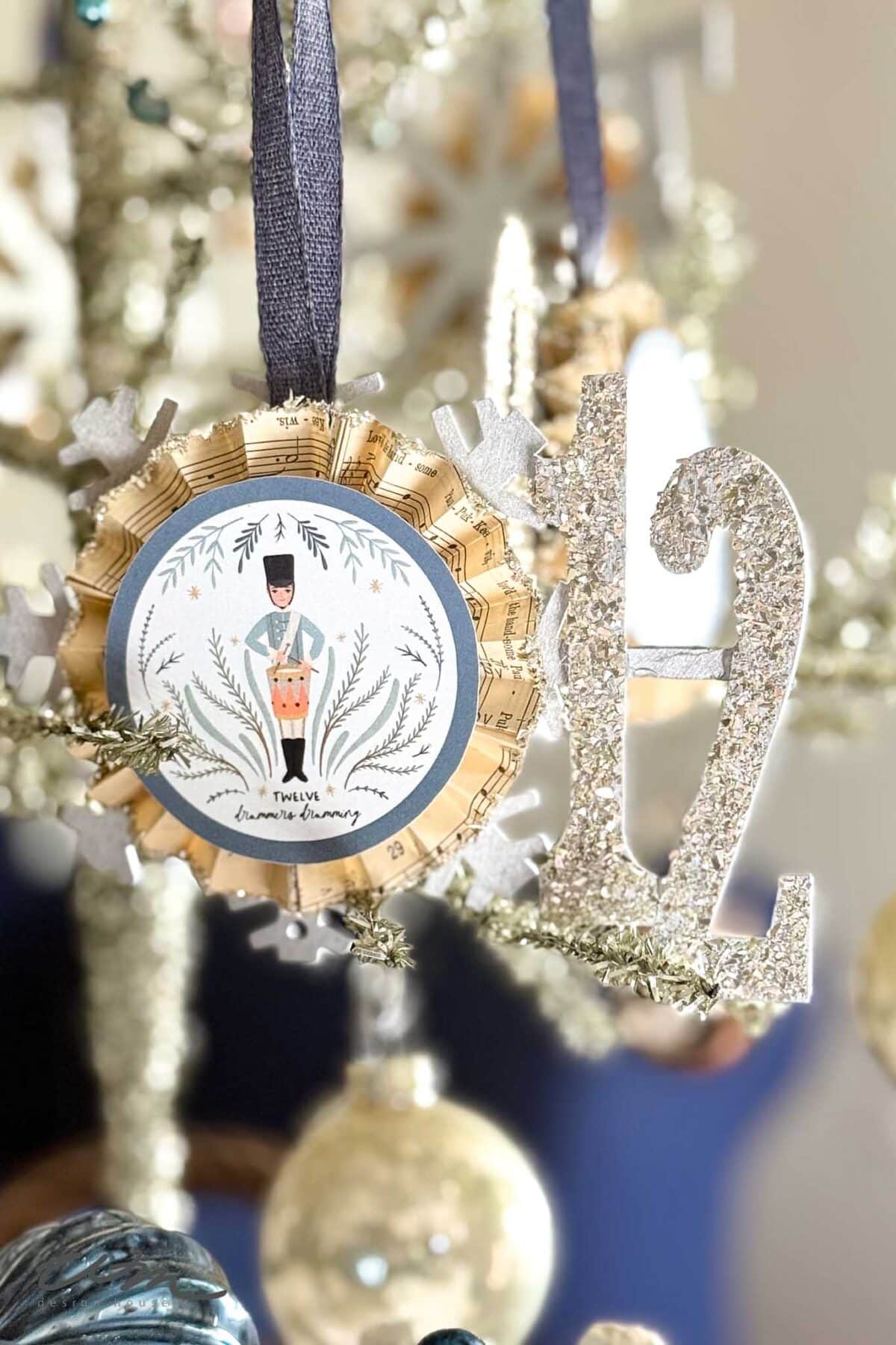 A close-up of a Christmas tree ornament with a glittery number "2" and a round medallion featuring a nutcracker illustration, hanging on the tree with blurred ornaments in the background.