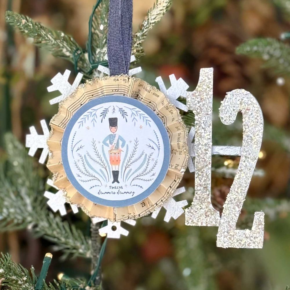 Close-up of a DIY 12 Days of Christmas ornament shaped like a snowflake, featuring a drummer and the words "Twelve Drummers Drumming." Next to it is a glittery number 12 decoration, both hanging on a Christmas tree.