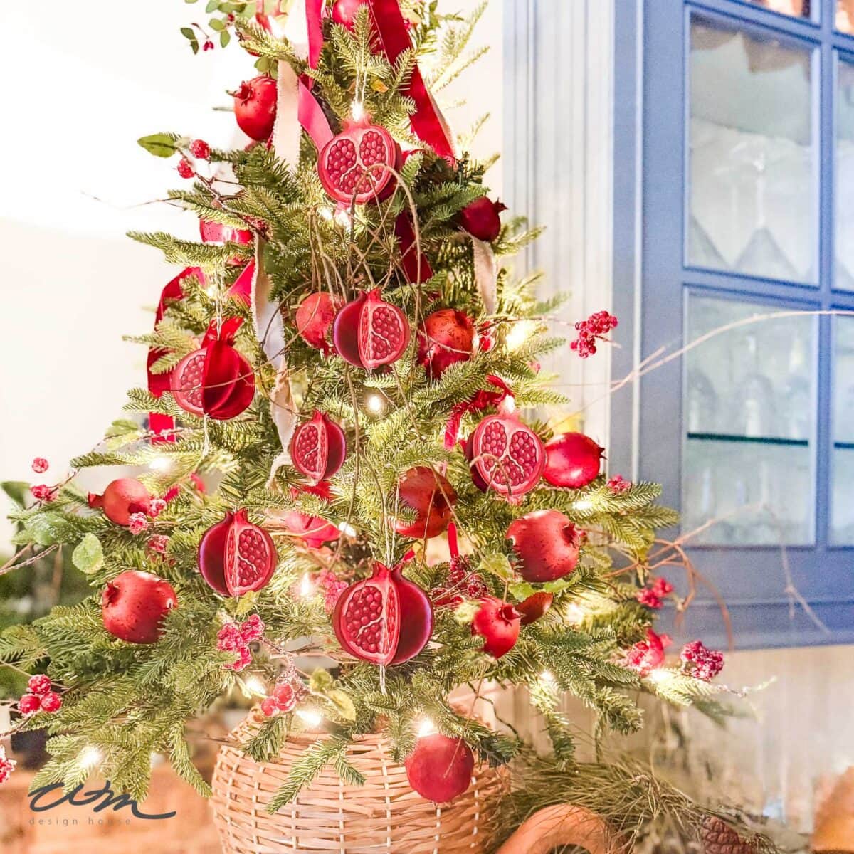 A pomegranate Christmas tree adorned with fairy lights and red ribbons sits in a woven basket near a blue cabinet, creating a festive and vibrant holiday display.