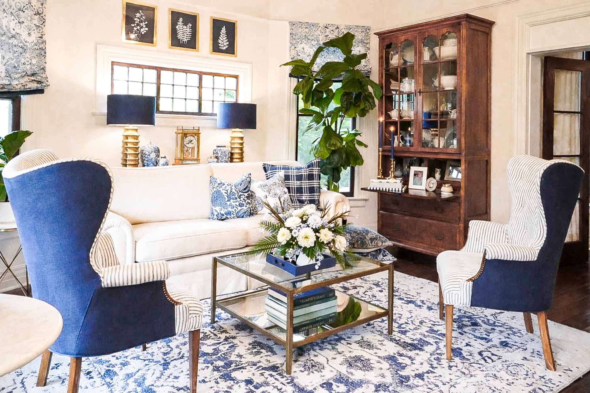 A cozy living room perfect for a winter home reset, featuring a white sofa, two blue armchairs, a glass coffee table with a floral arrangement, patterned rug, large indoor plant, and a wooden display cabinet against the wall.