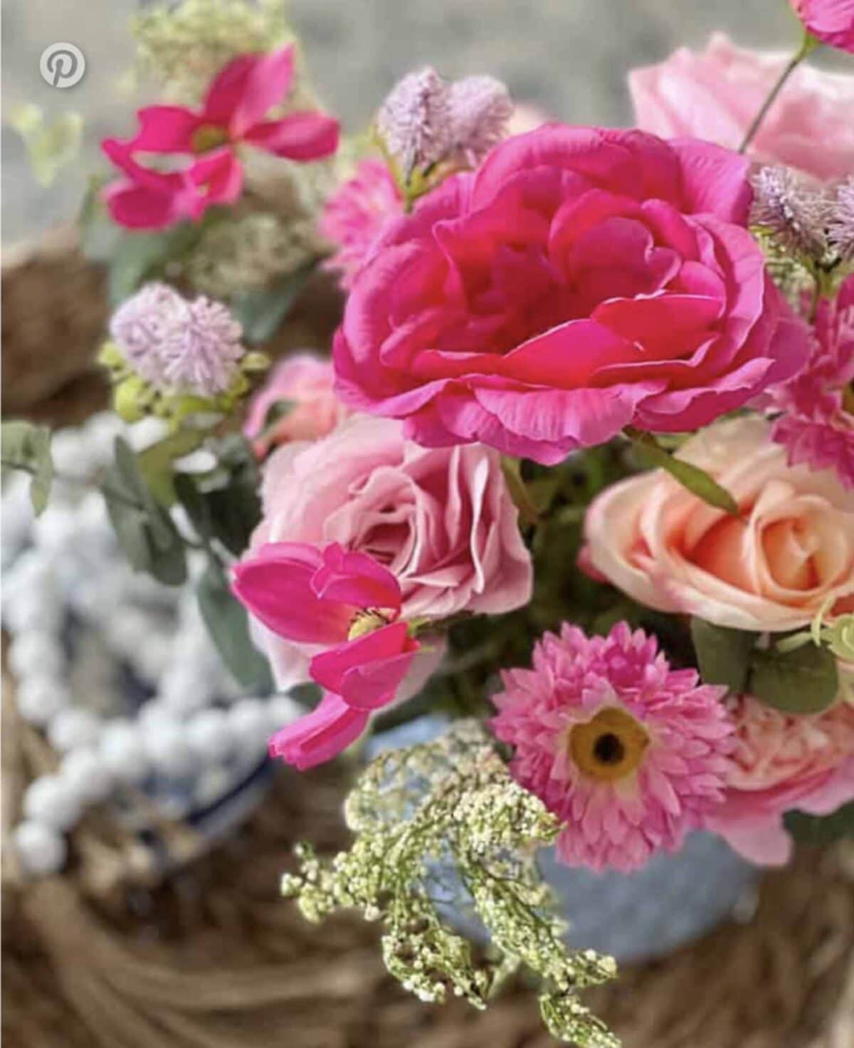 A close-up of a vibrant flower arrangement featuring pink roses, bright pink ranunculus, and peach flowers in a blue vase, with a blurred background and decorative white beads to the side.