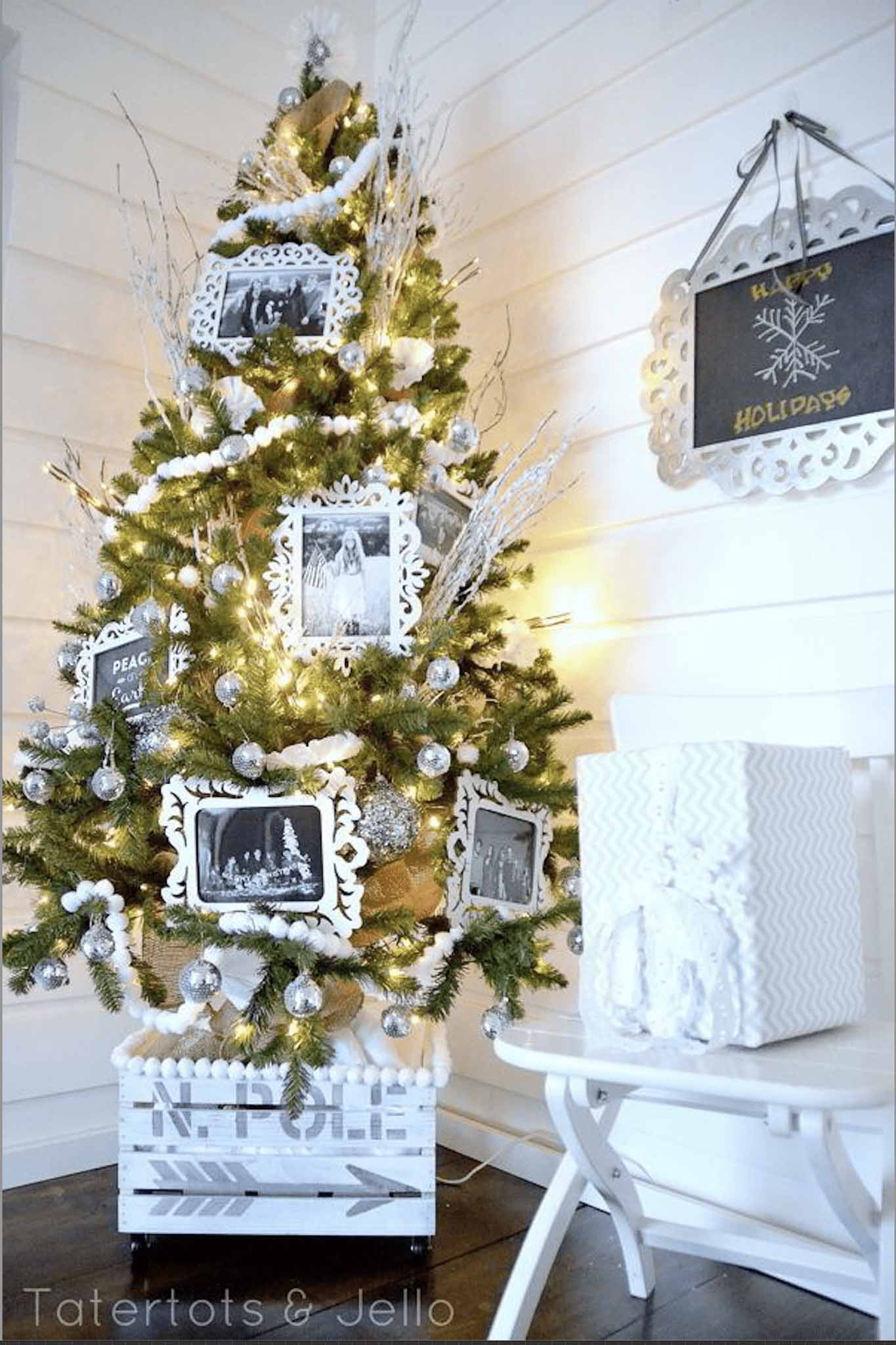 A Christmas tree decorated with white ornaments, framed black-and-white photos, and lights stands in a white room next to a chalkboard sign and a large, white gift box tied with a ribbon.