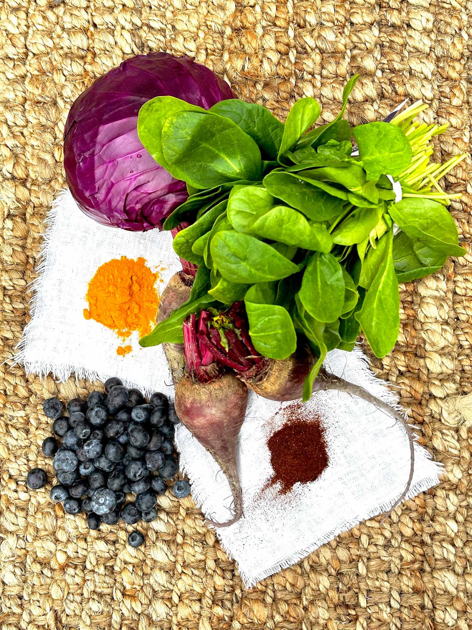 A bunch of fresh spinach, purple cabbage, blueberries, two beets, ground turmeric, and ground chili powder arranged on a woven surface with pieces of white cloth.