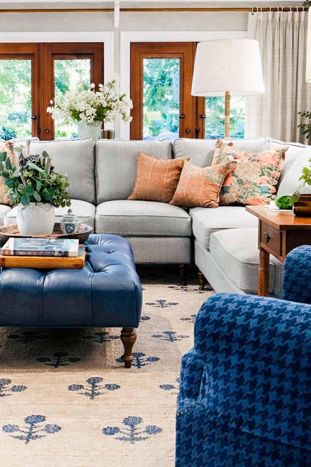 A cozy living room perfect for a winter home reset, featuring a light gray sectional sofa, patterned throw pillows, a blue tufted ottoman, blue armchair, wooden side table, large windows, and floral arrangements on the tables.