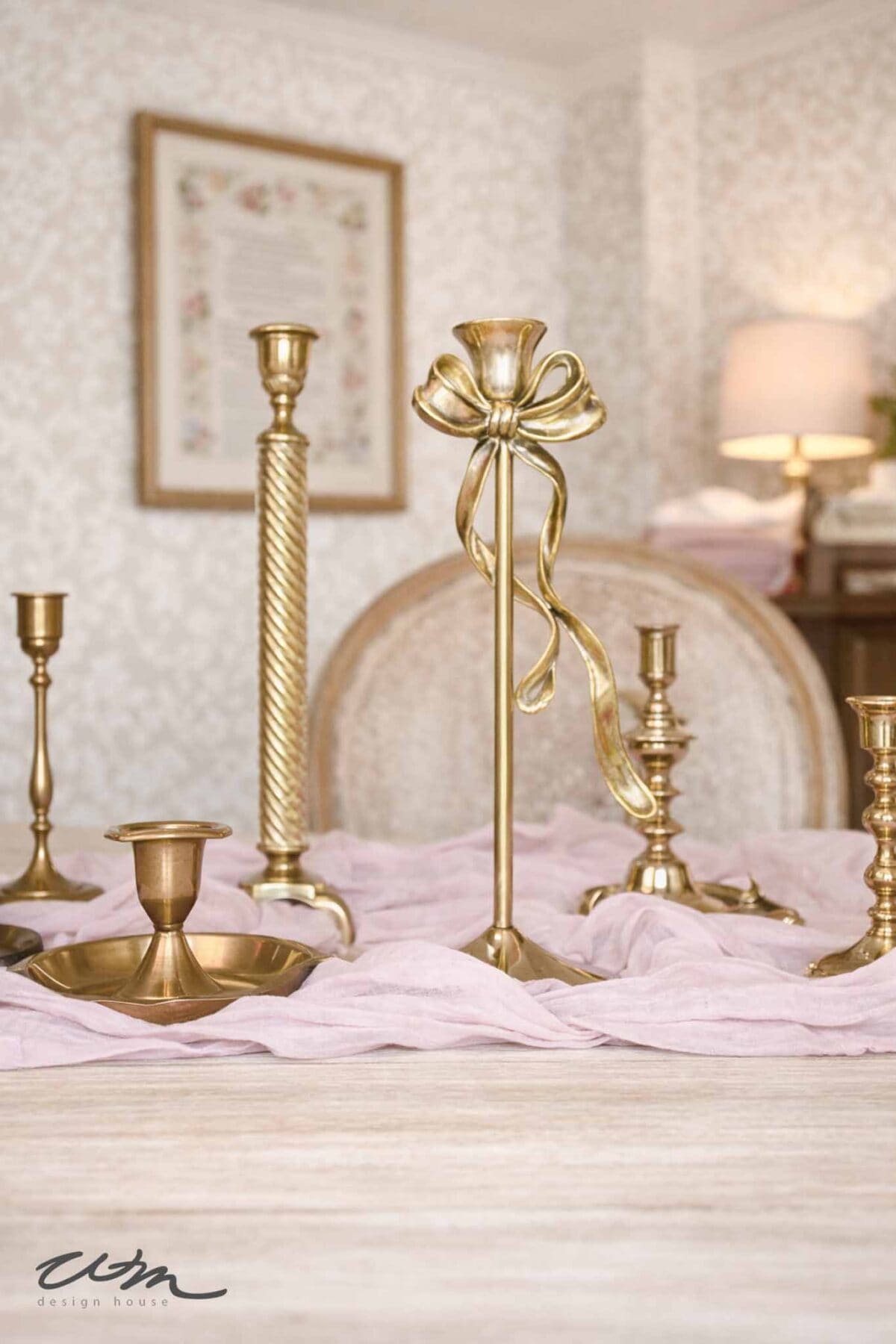 A collection of elegant gold candlesticks, including one with a decorative bow, arranged for a Valentine’s party for grandkids on a wooden table draped with light pink cloth. A soft, neutral room with floral wallpaper is in the background.
