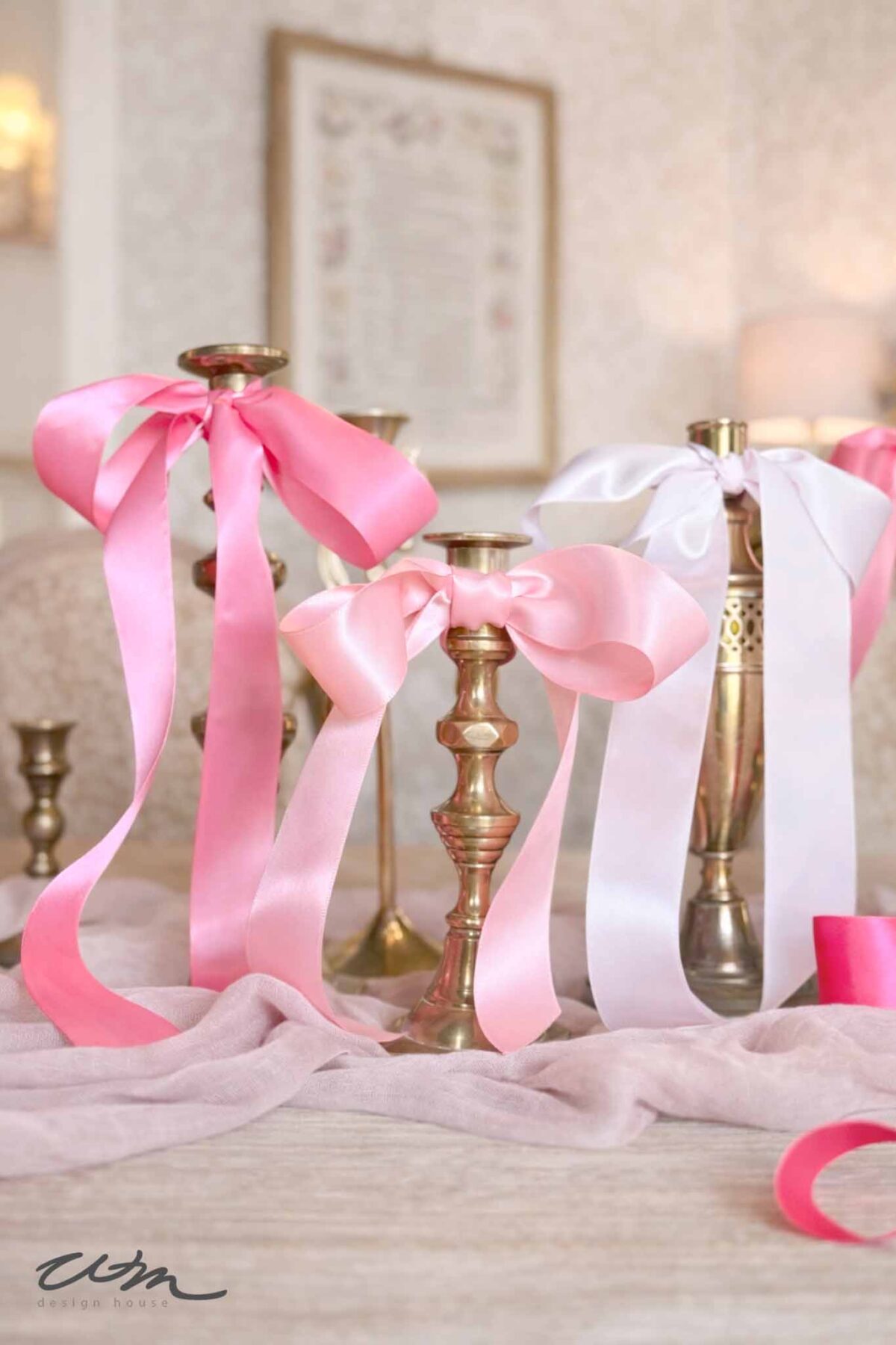 Three brass candlesticks decorated with pink and white satin ribbons sit on a light pink fabric-covered table, creating a charming centerpiece perfect for a Valentine's party for grandkids. The softly blurred background displays framed wall art.