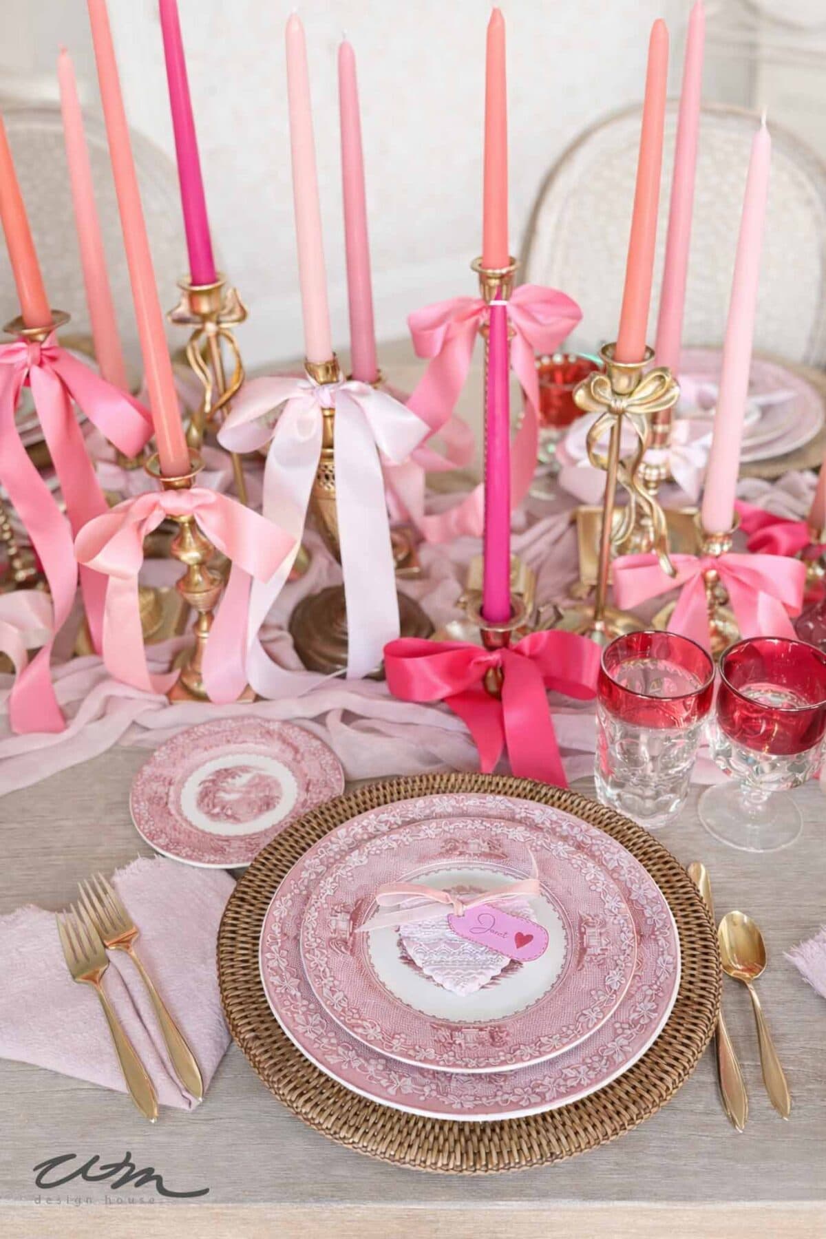 A romantic table setting perfect for a Valentine’s party for grandkids, with pink and red candles in gold holders, vintage-style pink plates, gold cutlery, pink napkins, heart-shaped “Love” card, and festive red-tinted glasses.