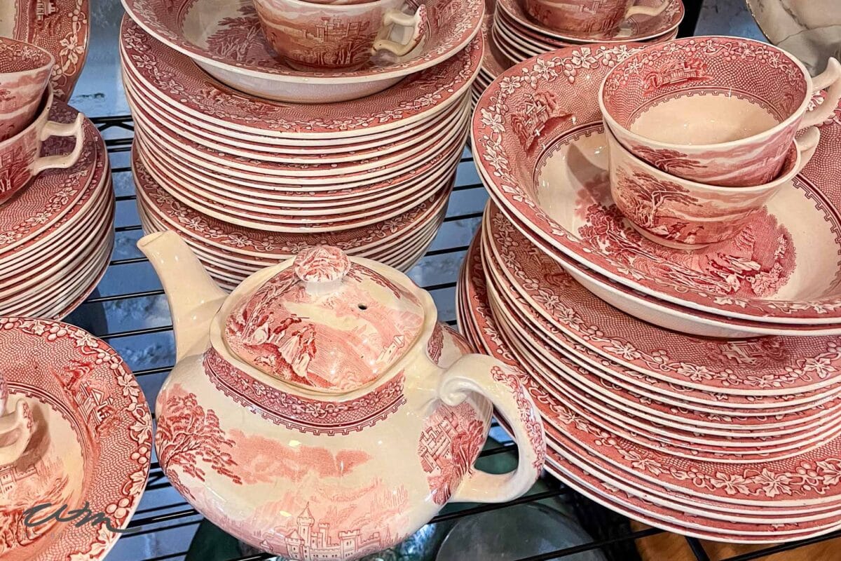 A collection of vintage red and white china dishes, perfect for a Valentine’s party for grandkids, featuring plates, cups, saucers, and a teapot with intricate pastoral and floral patterns, all arranged neatly on a metal shelf.