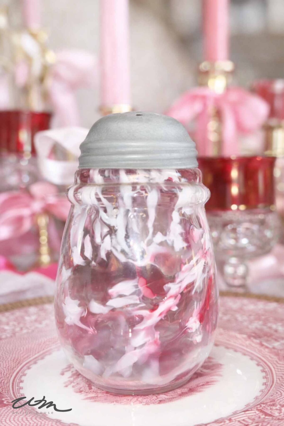 A vintage glass shaker with a silver metal lid sits on a pink and white decorative plate, perfect for a Valentine’s party for grandkids, with a blurred background featuring pink candles and ribbon accents.