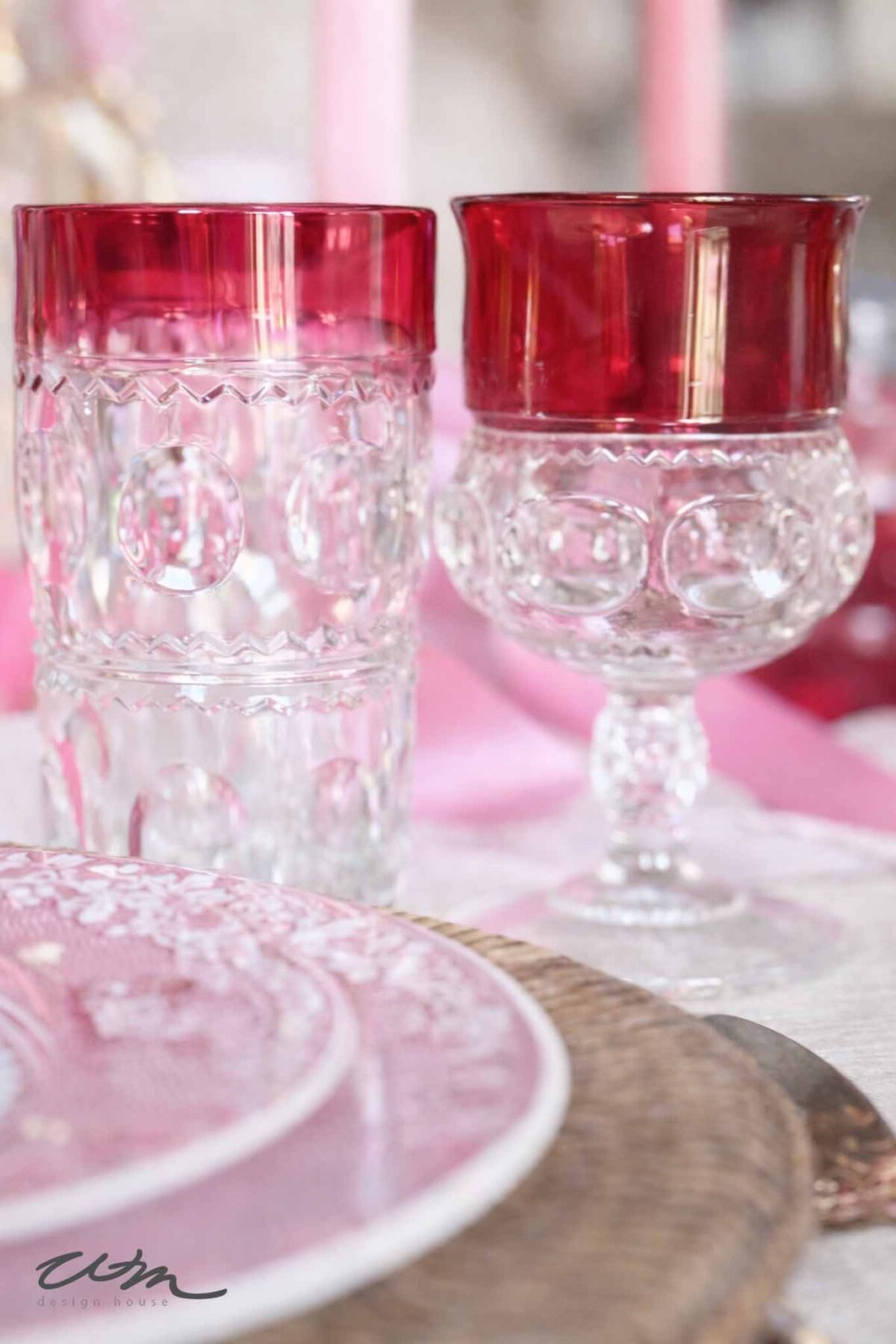 Two vintage-style glasses with ornate clear bases and deep red rims sit on a table set with pink-patterned plates, perfect for a Valentine’s party for grandkids. The scene has a soft, romantic ambiance.