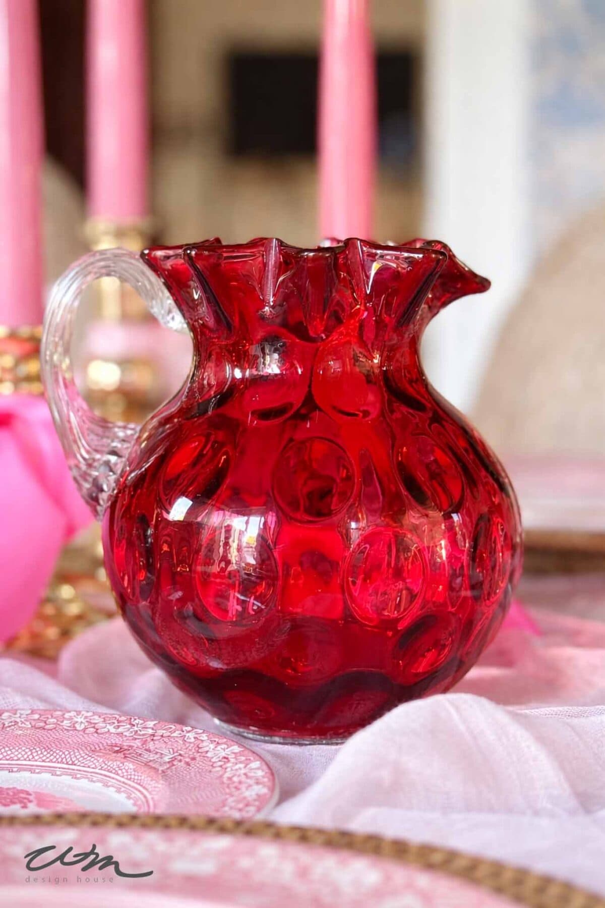 A vibrant red glass pitcher with a textured, bubble-like pattern and a clear handle sits on a pink and white table setting, perfect for a Valentine’s party for grandkids, with pink candles and plates in the background.