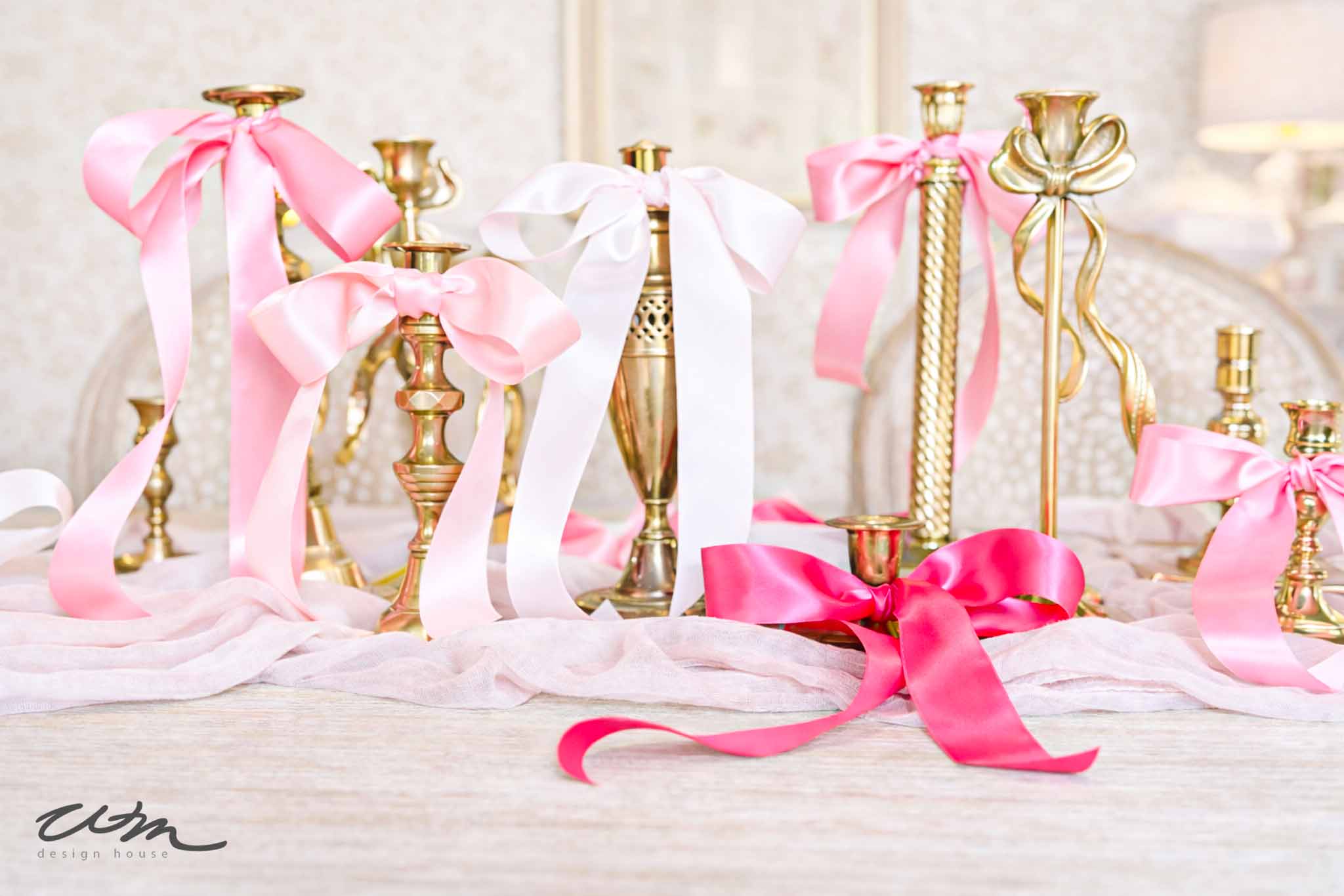 A collection of gold candlesticks decorated with large pink and white satin bows is arranged on a table draped with soft pink fabric—perfect for a Valentine’s party for grandkids in a light, elegant room.