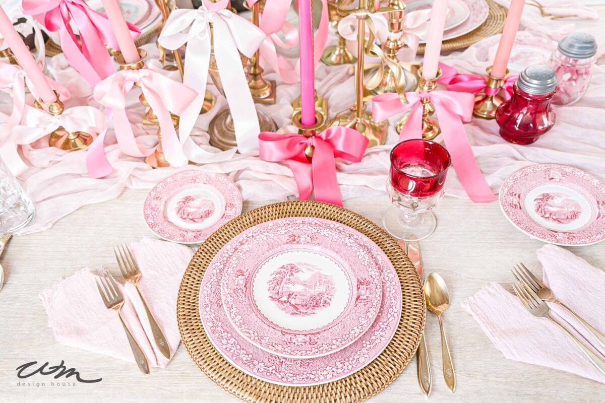 A table set with pink and white vintage china, gold cutlery, a pink napkin, a red wine glass, and gold candleholders with pink ribbons and candles creates an elegant, romantic atmosphere—perfect for a Valentine’s party for grandkids.