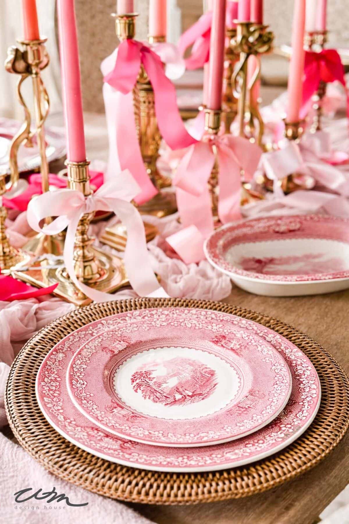 A table setting with pink and white patterned plates on woven chargers, surrounded by brass candlestick holders with pink taper candles and ribbon bows, creates a romantic, elegant atmosphere—perfect for a Valentine’s party for grandkids.