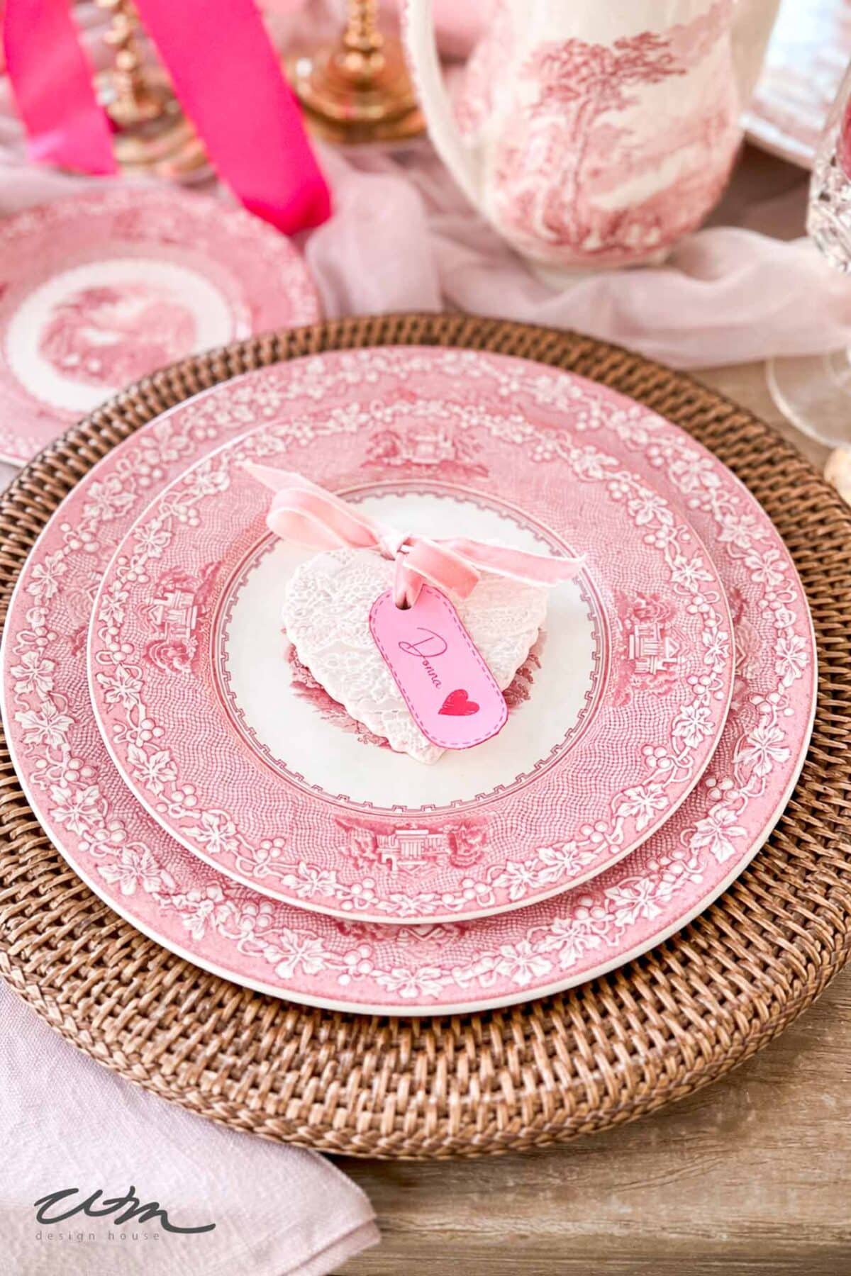 A place setting with pink and white patterned plates on a woven charger, topped with a heart-shaped lace ornament and a pink “Darling” tag, perfect for creating a romantic, vintage vibe at a Valentine’s party for grandkids.