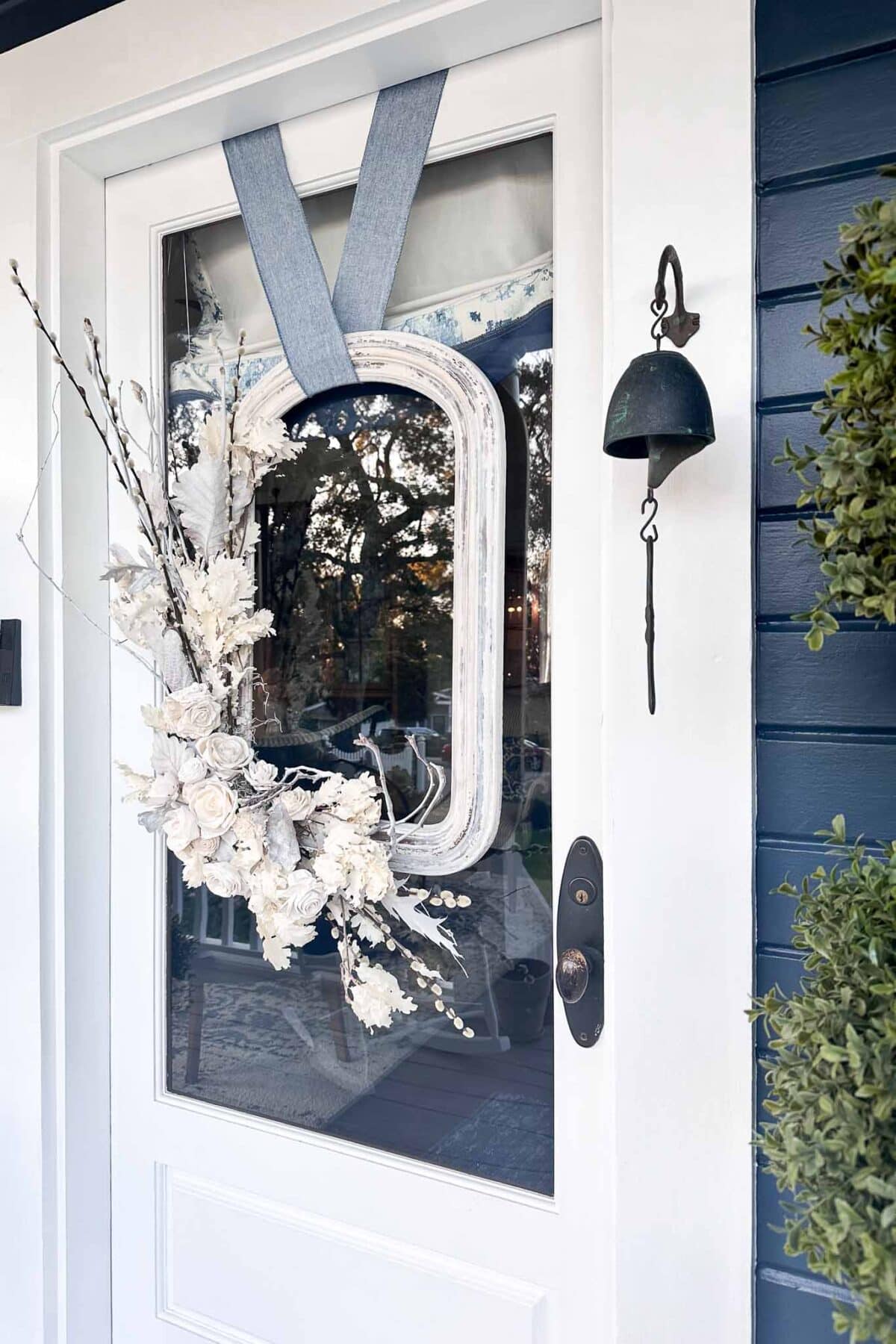 A glass-paneled white door with a unique rectangular floral wreath made of white flowers and branches, hanging by a blue ribbon. A black doorbell and a metal bell are mounted on the dark blue exterior wall beside the door.