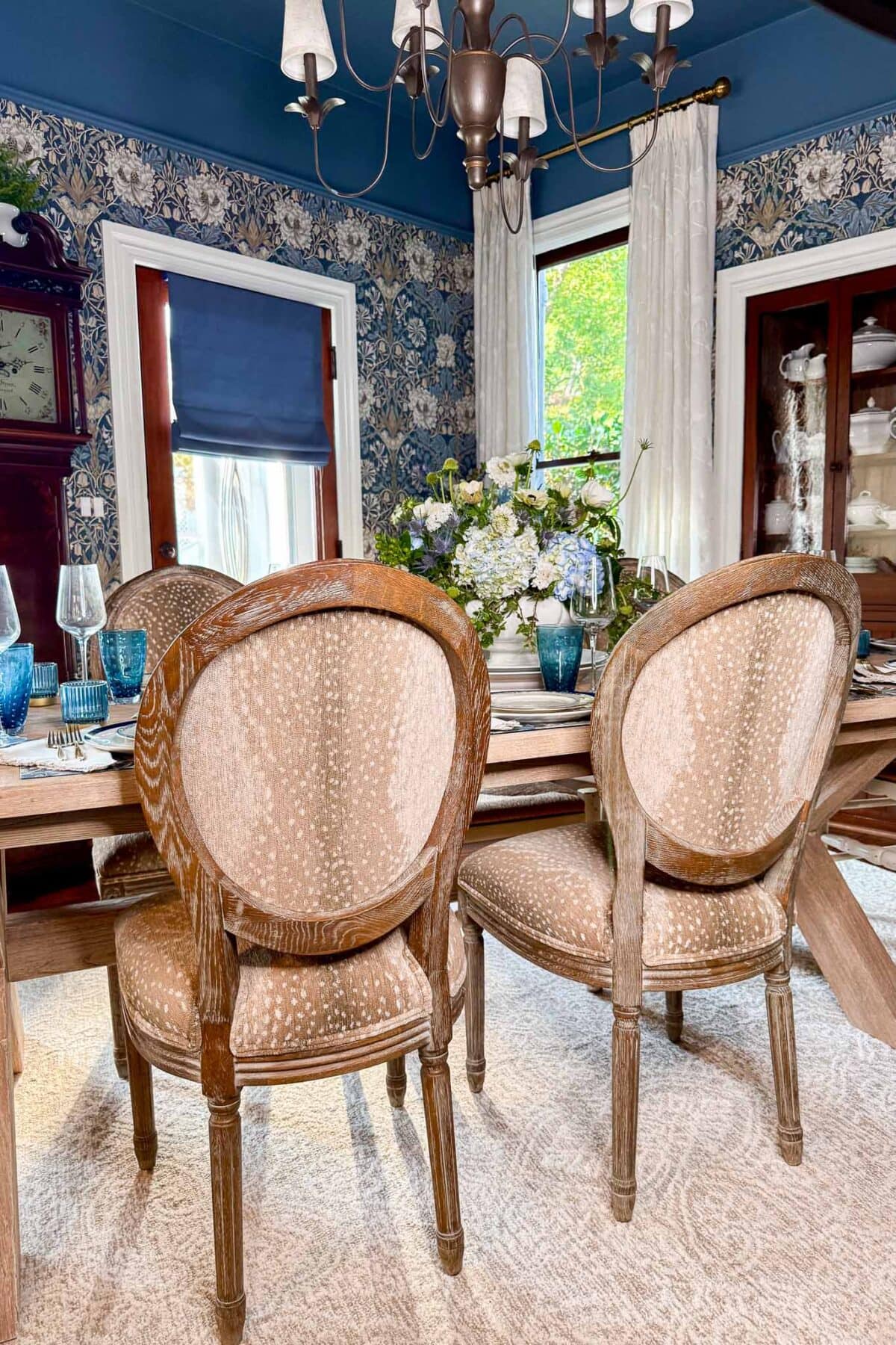 A dining room with oval-backed wooden chairs, a set table with blue glasses, a floral centerpiece, wallpaper with a floral pattern, and large windows with cream curtains and blue Roman shades.